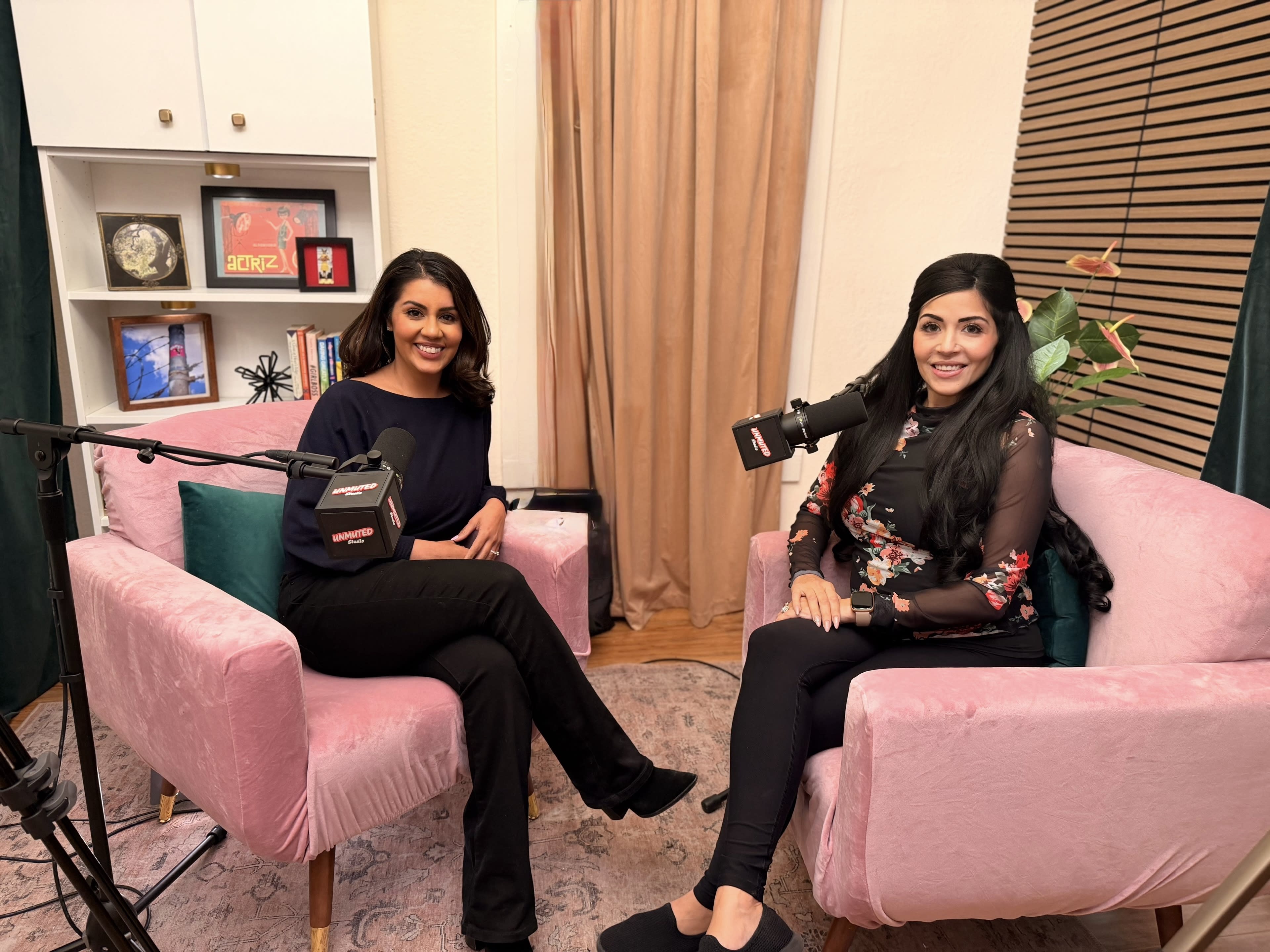 Two women sit in pink chairs in a well-decorated studio, each with a microphone in front of them, engaged in conversation.