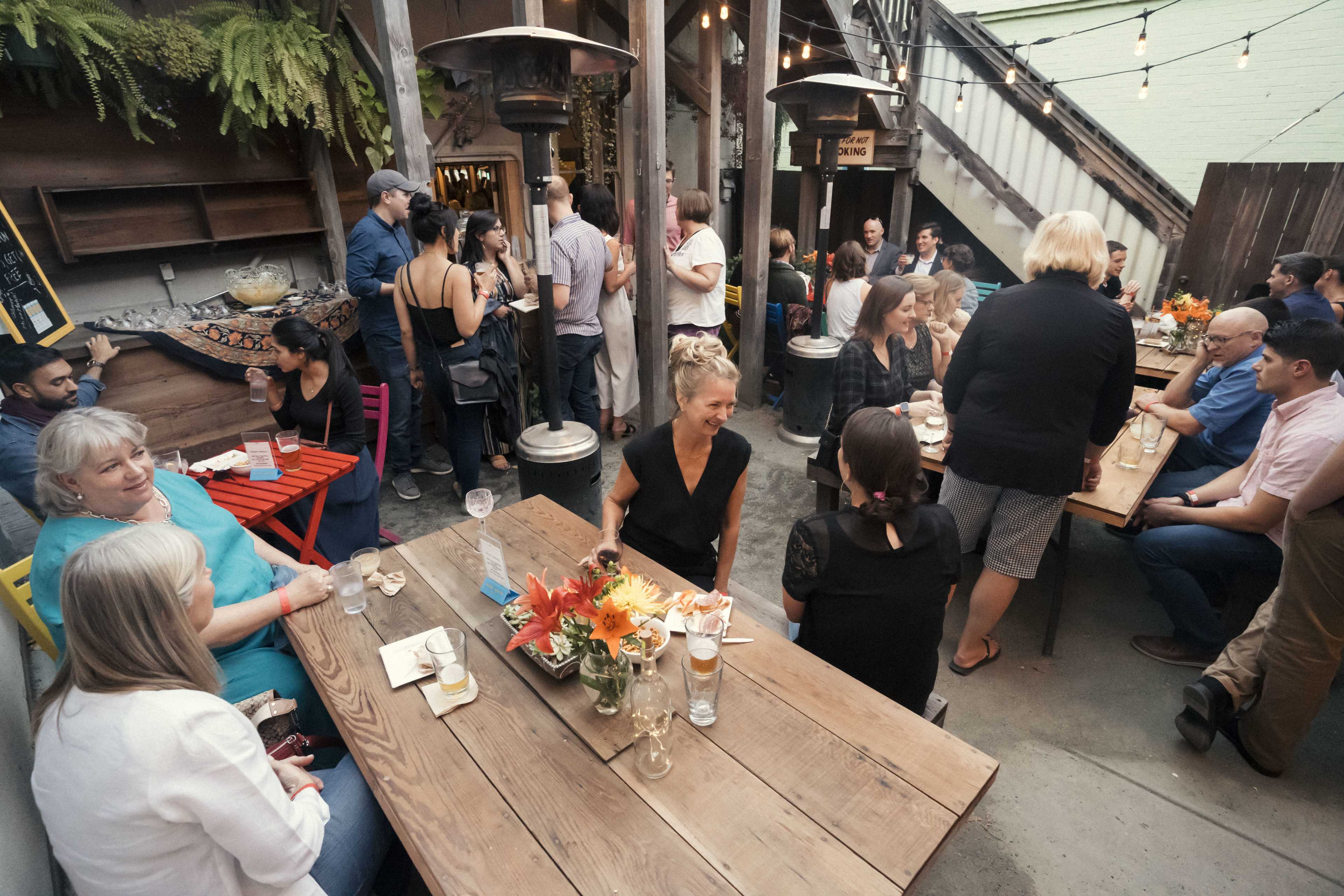 A group of people socializes in a cozy outdoor setting with wooden tables, vibrant flowers, and dim string lights.