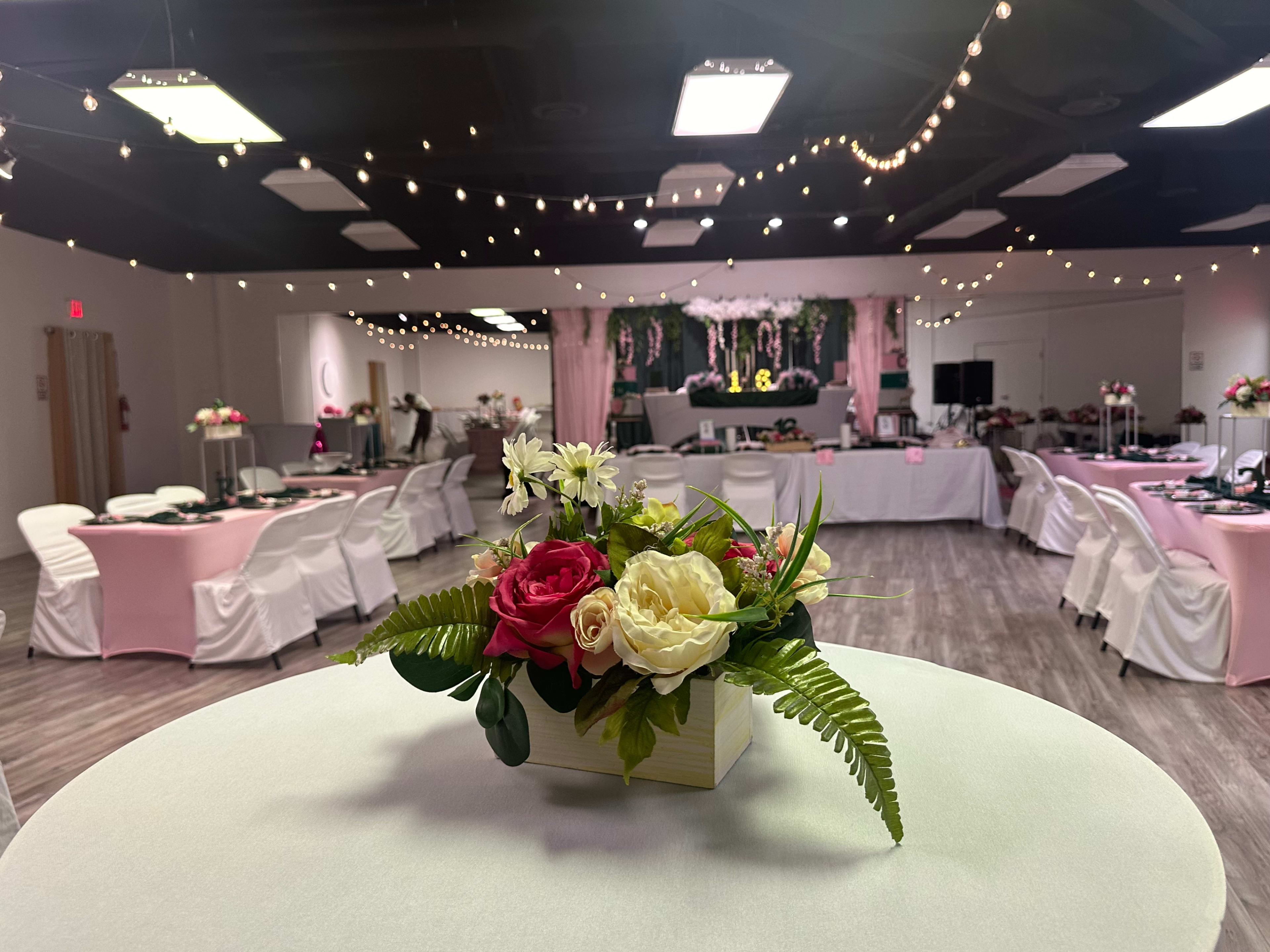 The image shows a banquet hall decorated for an event, featuring floral centerpieces on white tables and string lights overhead.