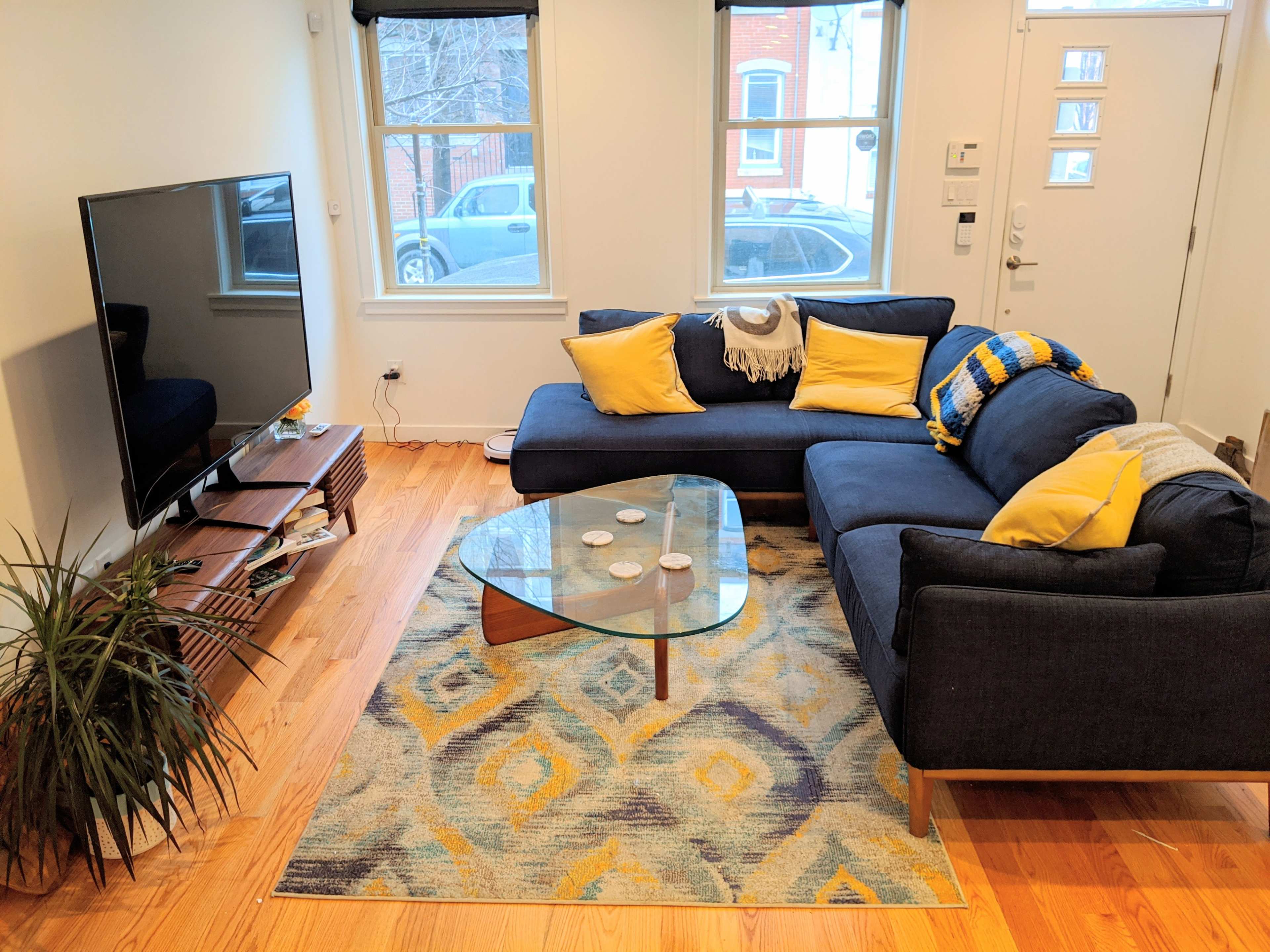 A living room with a dark blue sectional sofa, a glass coffee table, a television mounted on the wall, and a patterned area rug on a hardwood floor.