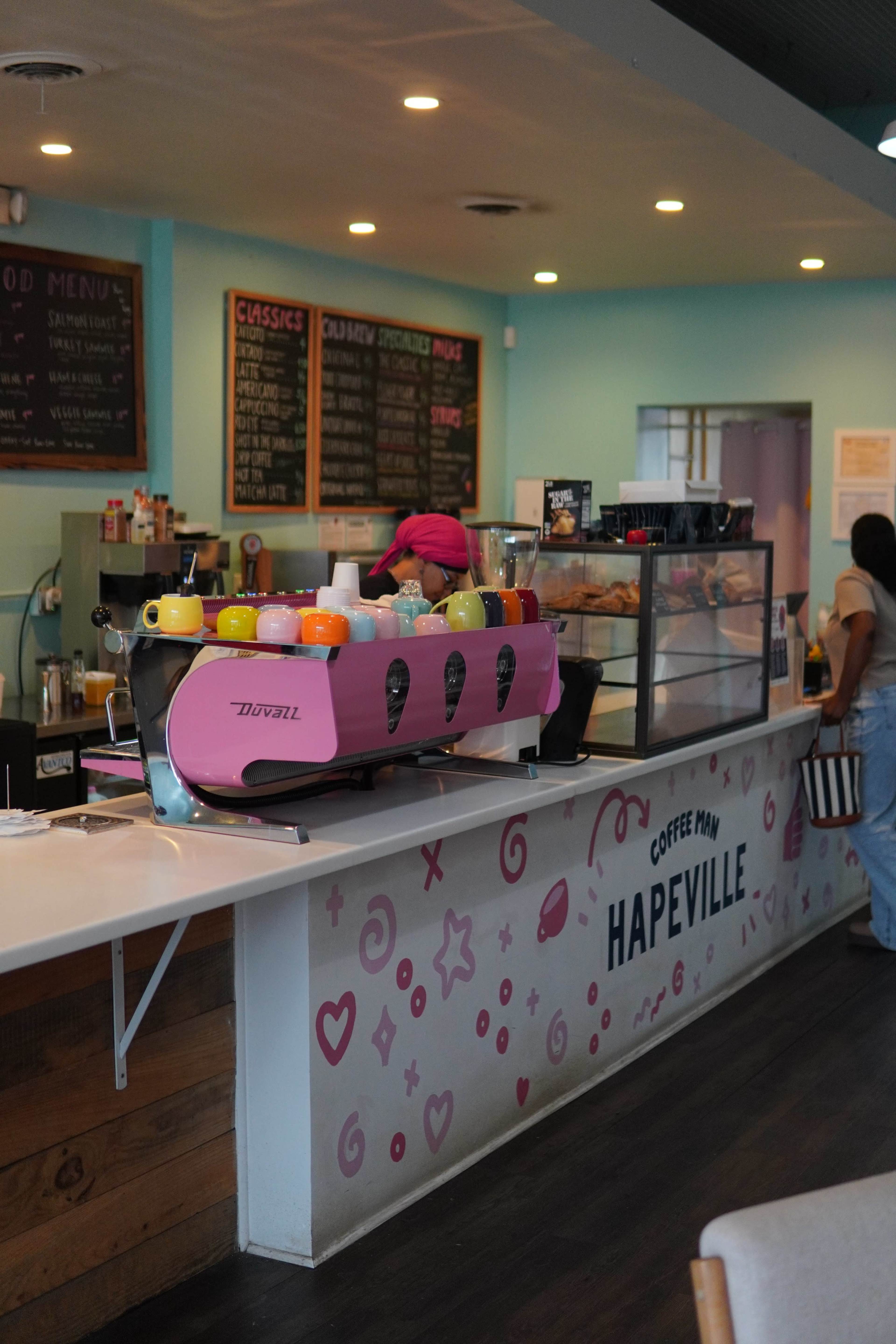 A pink espresso machine sits on the counter of a café displaying a colorful array of drinks, while a barista prepares beverages in the background.