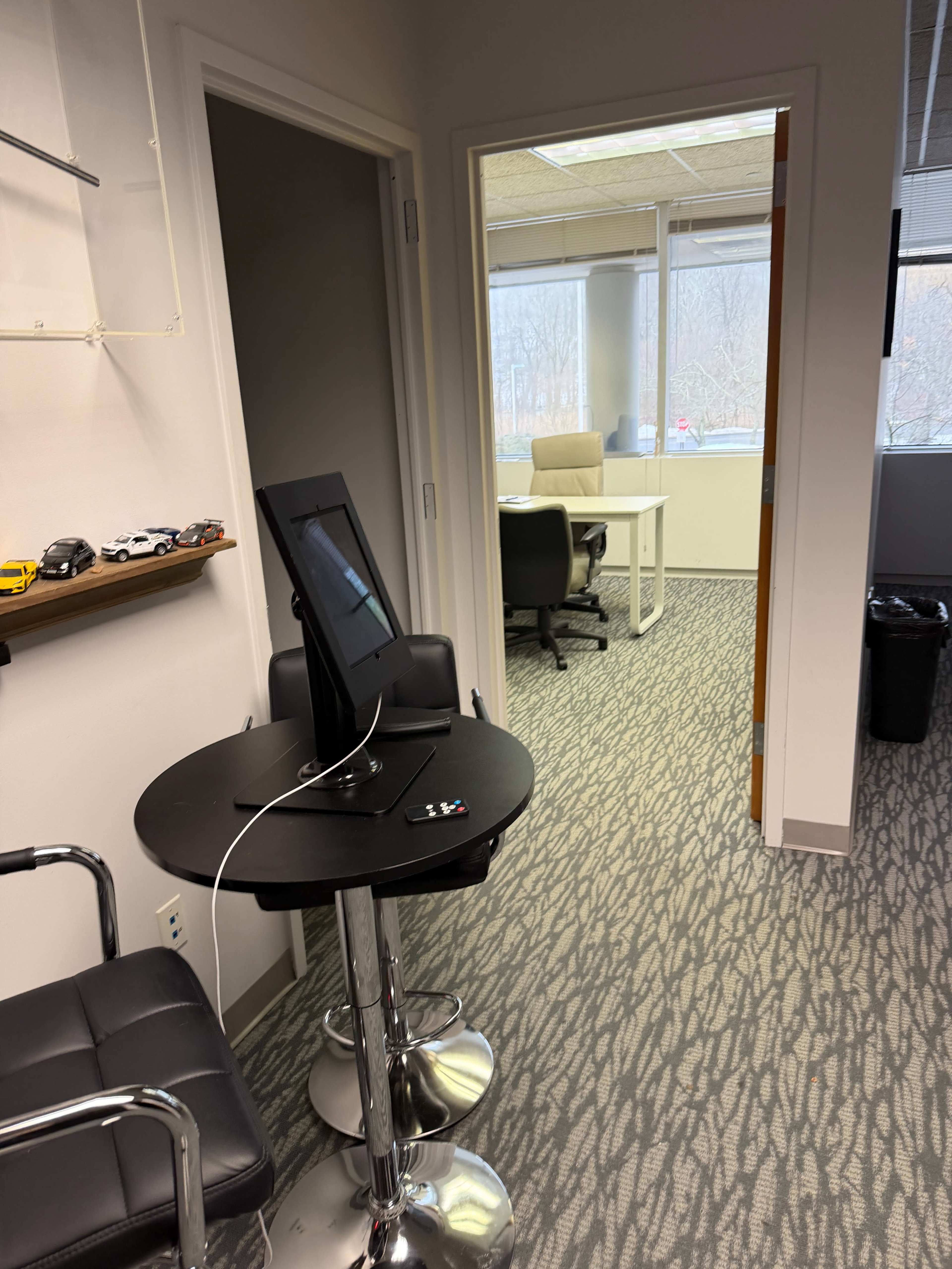 The image shows an office corridor with a high table and monitor on the left, leading to a bright office area with a desk and chairs visible through a doorway.