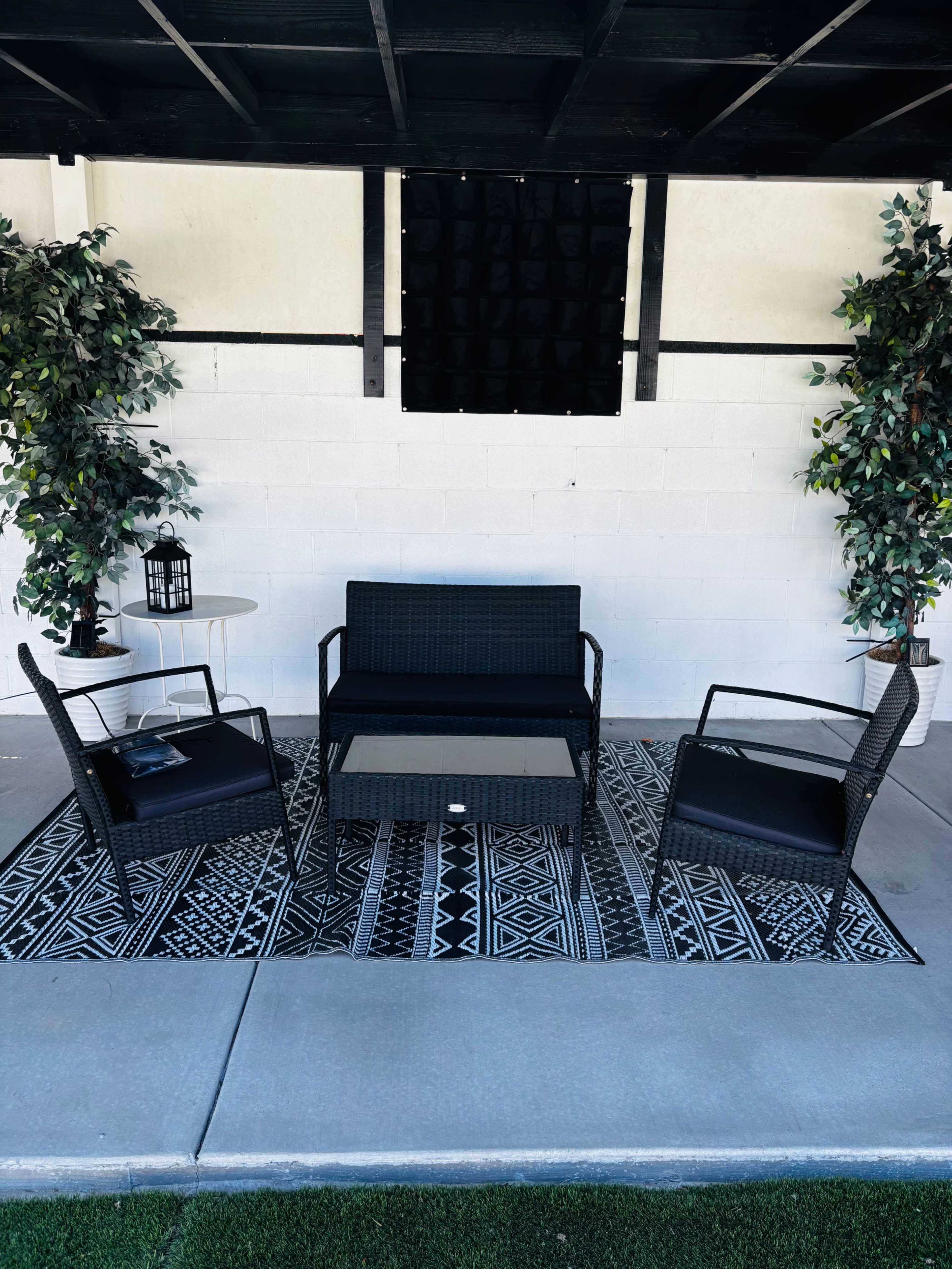 The image shows a small outdoor seating area with a black wicker sofa, two matching chairs, a coffee table, and decorative artificial plants arranged on a patterned rug.