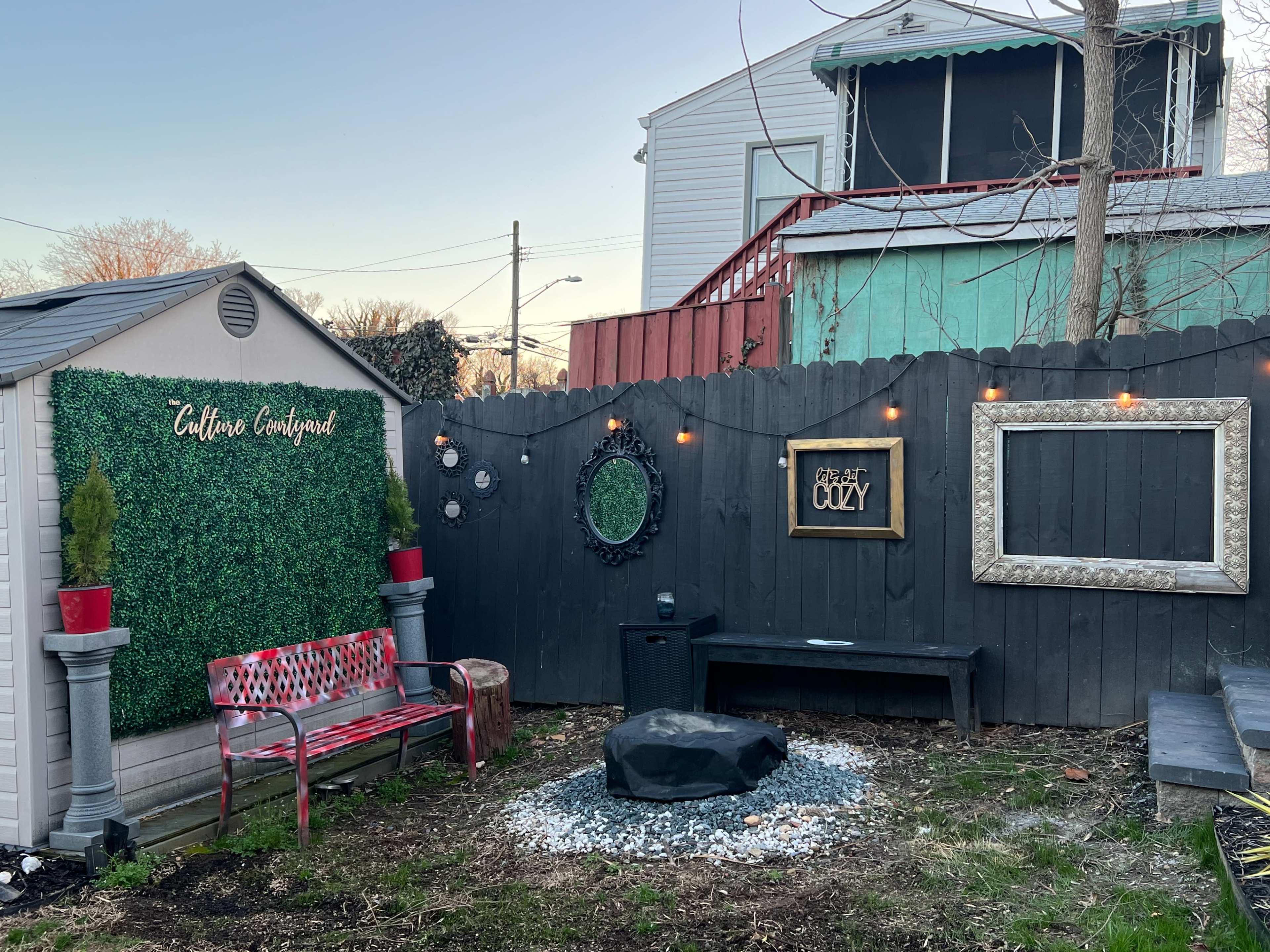 The image shows a small outdoor space featuring a red bench, a fire pit surrounded by gravel, decorative mirrors, and a green wall with the words "Caffeine Confined."