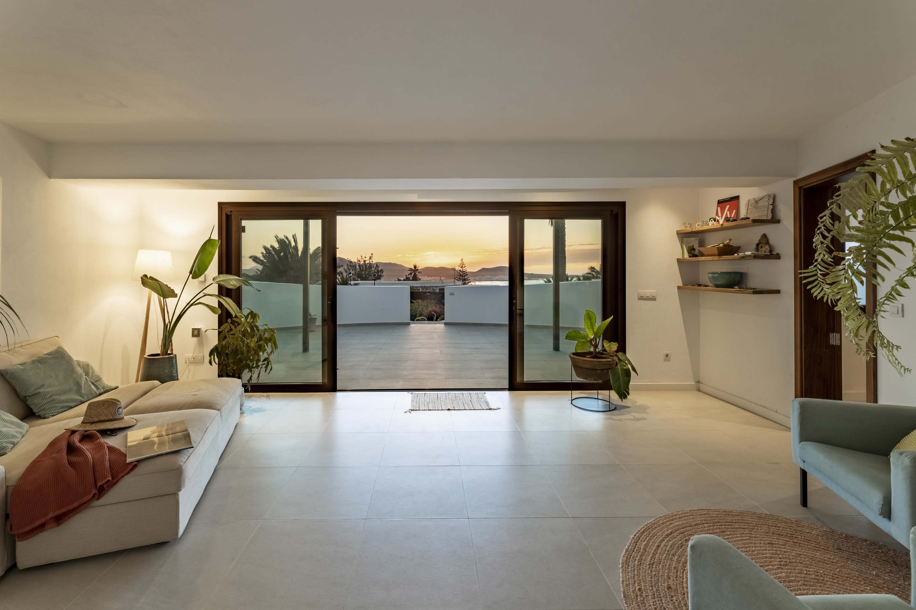 A living room features a large glass sliding door opening to a patio with a sunset view, accompanied by a sofa, plants, and decorative elements.