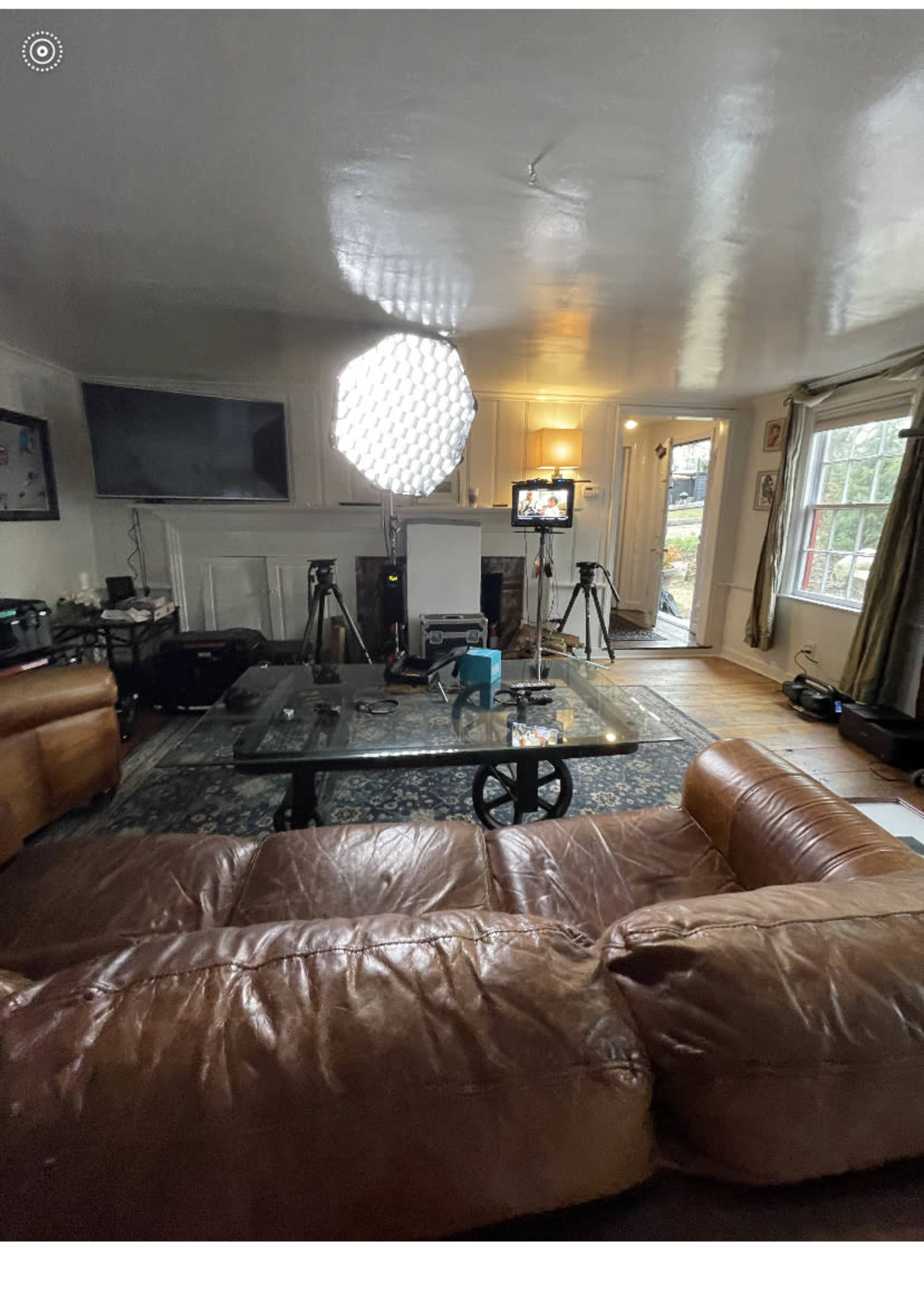 The image shows a living room scene featuring a large light source, cameras, and a coffee table on a rug, with a television mounted on the wall and a door leading outside.