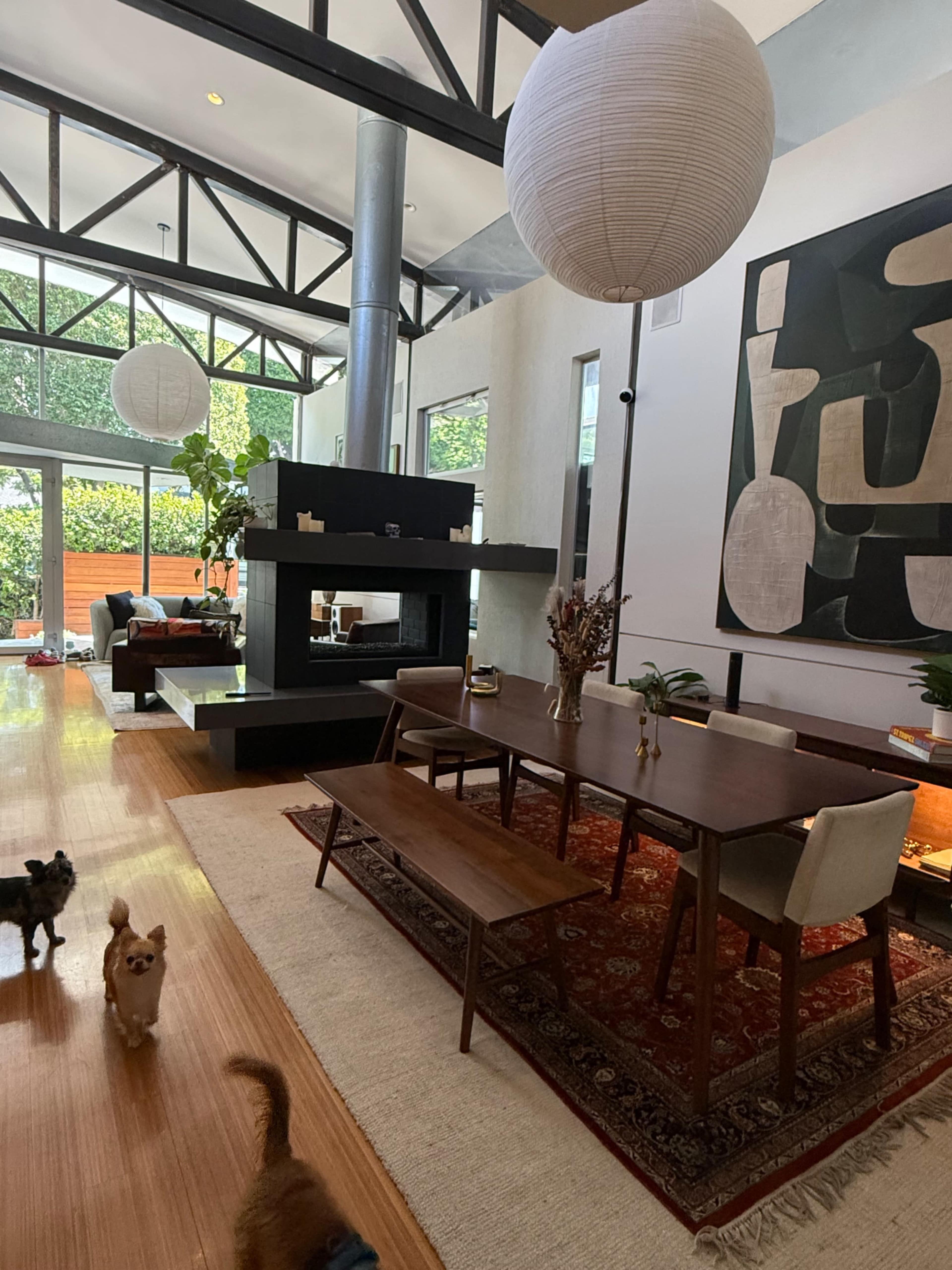 The image shows a modern interior space featuring a dining area with a wooden table, a large abstract painting on the wall, and a cozy seating area with natural light coming through large windows.