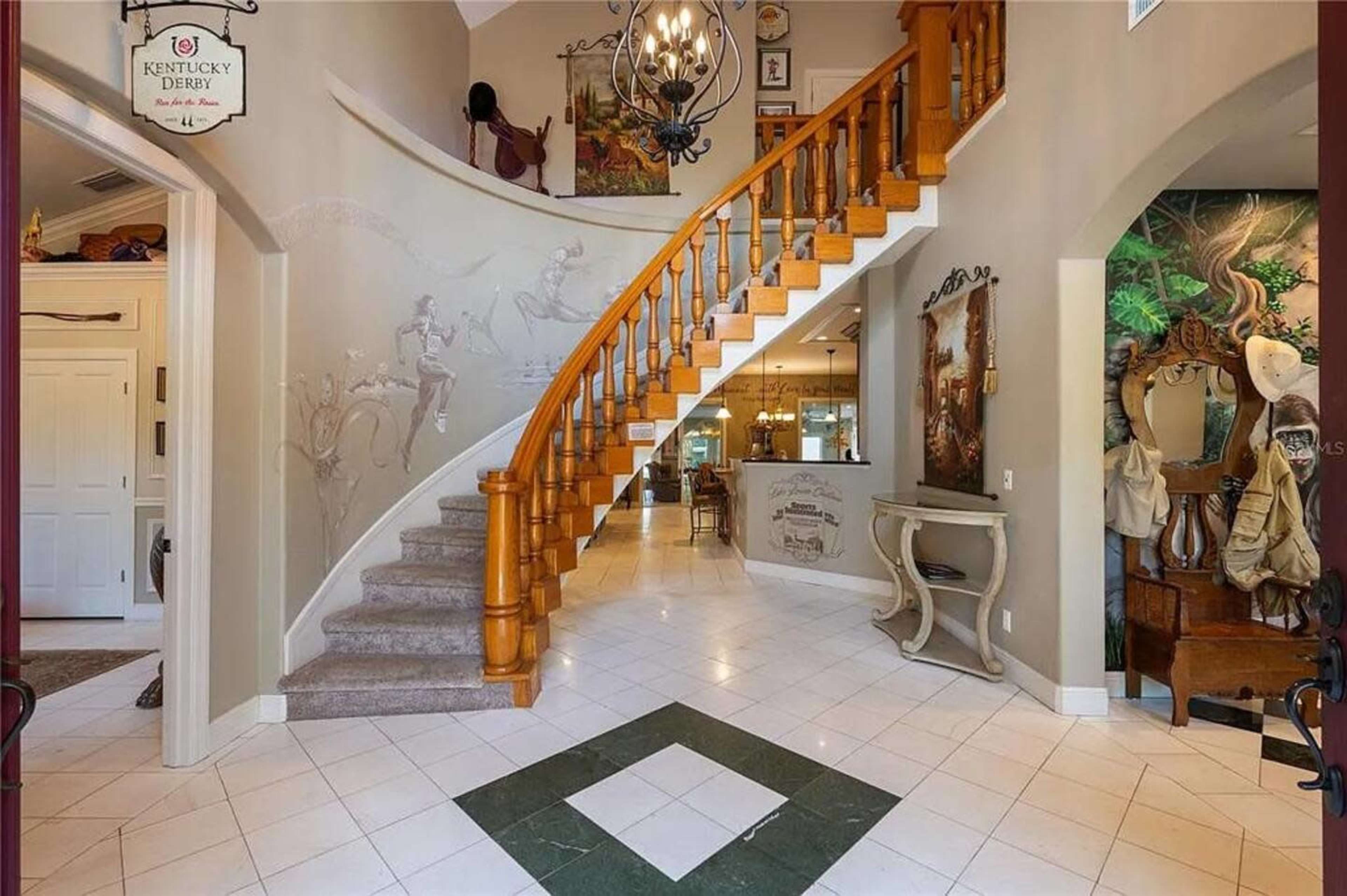 The image shows a spacious entryway featuring a sweeping wooden staircase, tiled flooring, and decorative art on the walls.