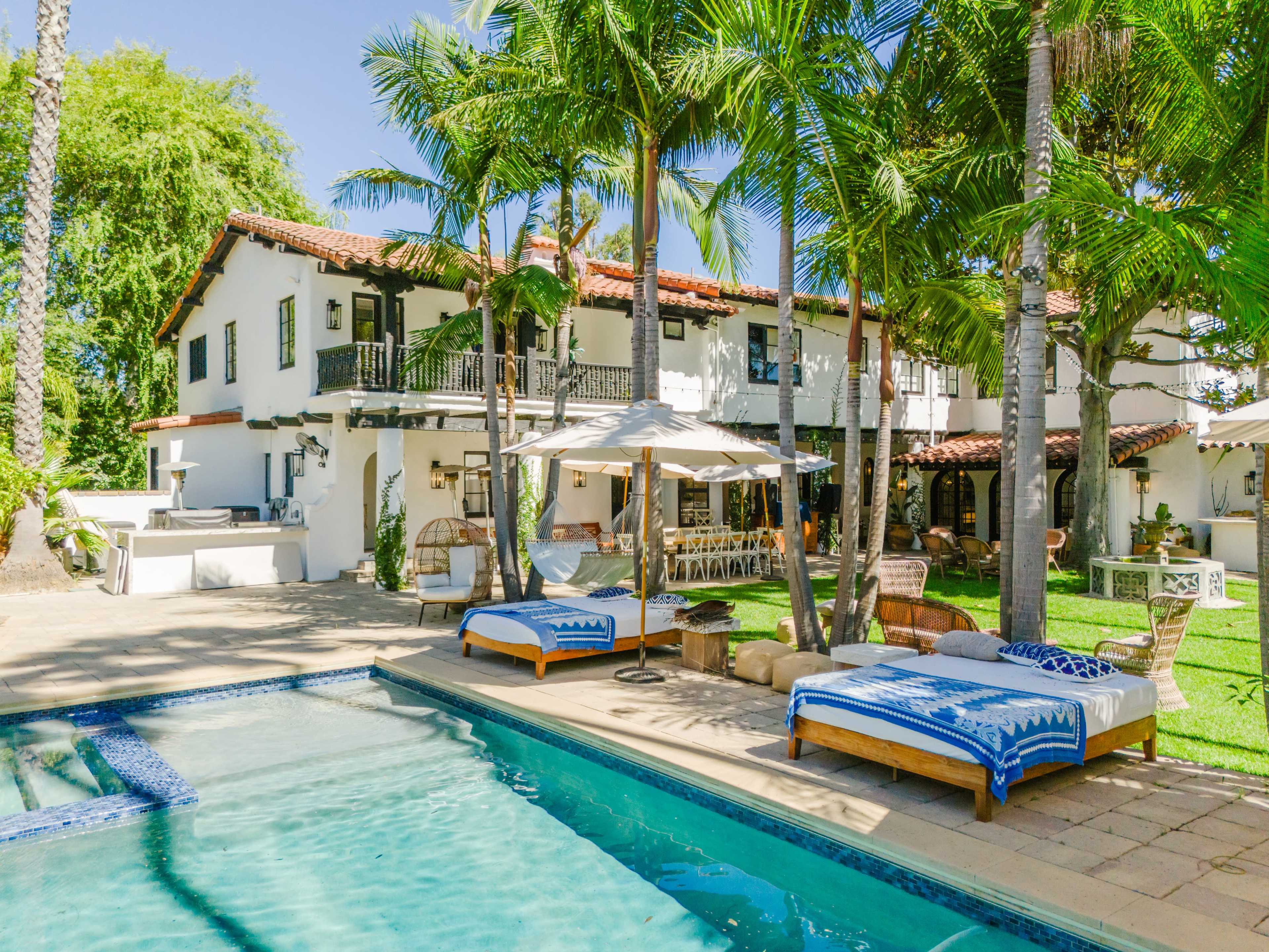 A tranquil backyard scene features a swimming pool surrounded by lounge chairs and palm trees, with a two-story white house in the background.