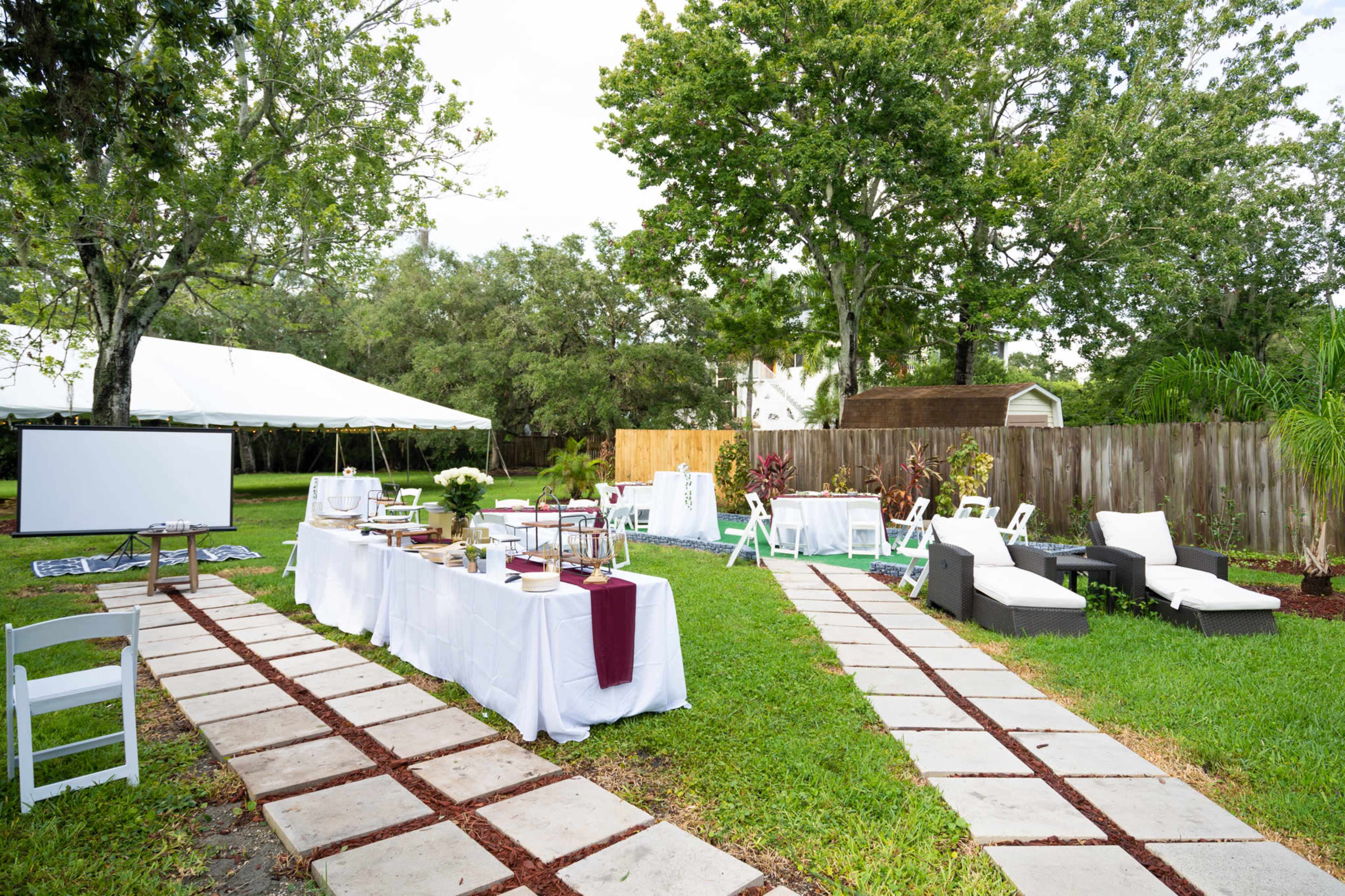 A backyard setup for an event, featuring tables with white tablecloths, chairs, and a tent in the background.