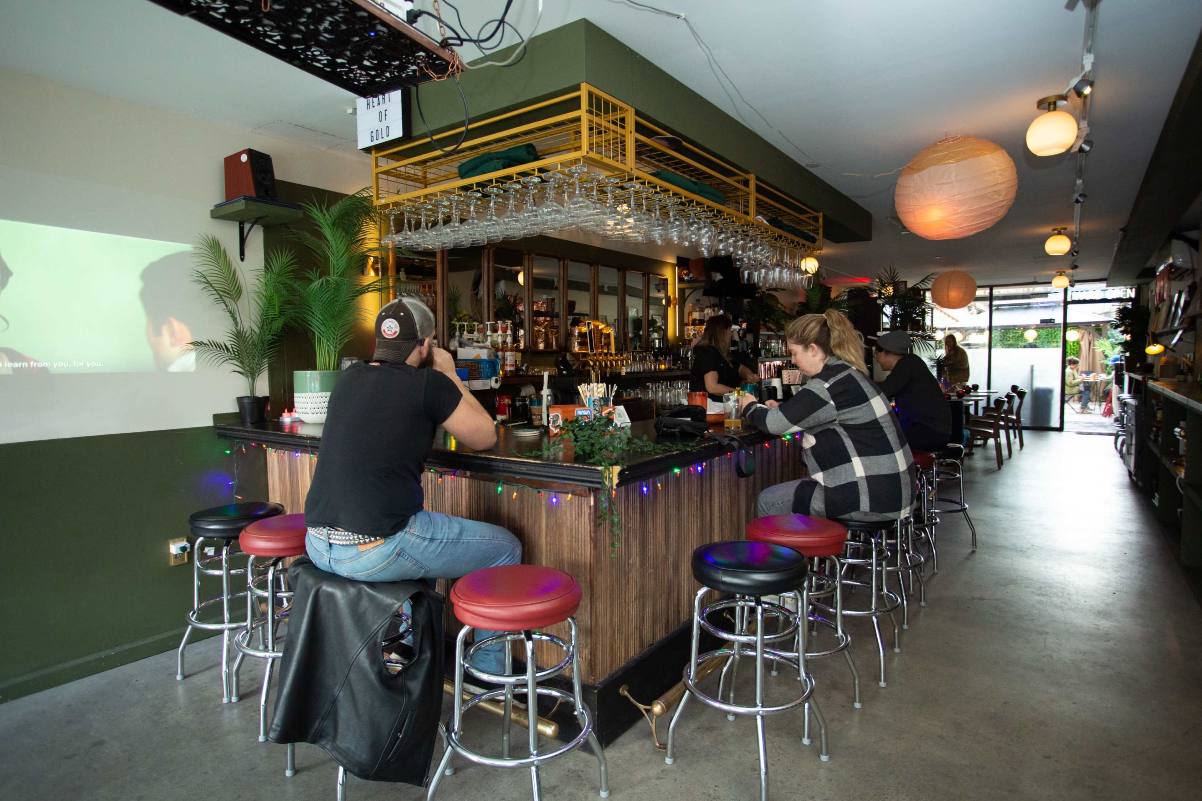 A bar scene features patrons seated at a long counter with a row of stools, surrounded by various plants and dim lighting.