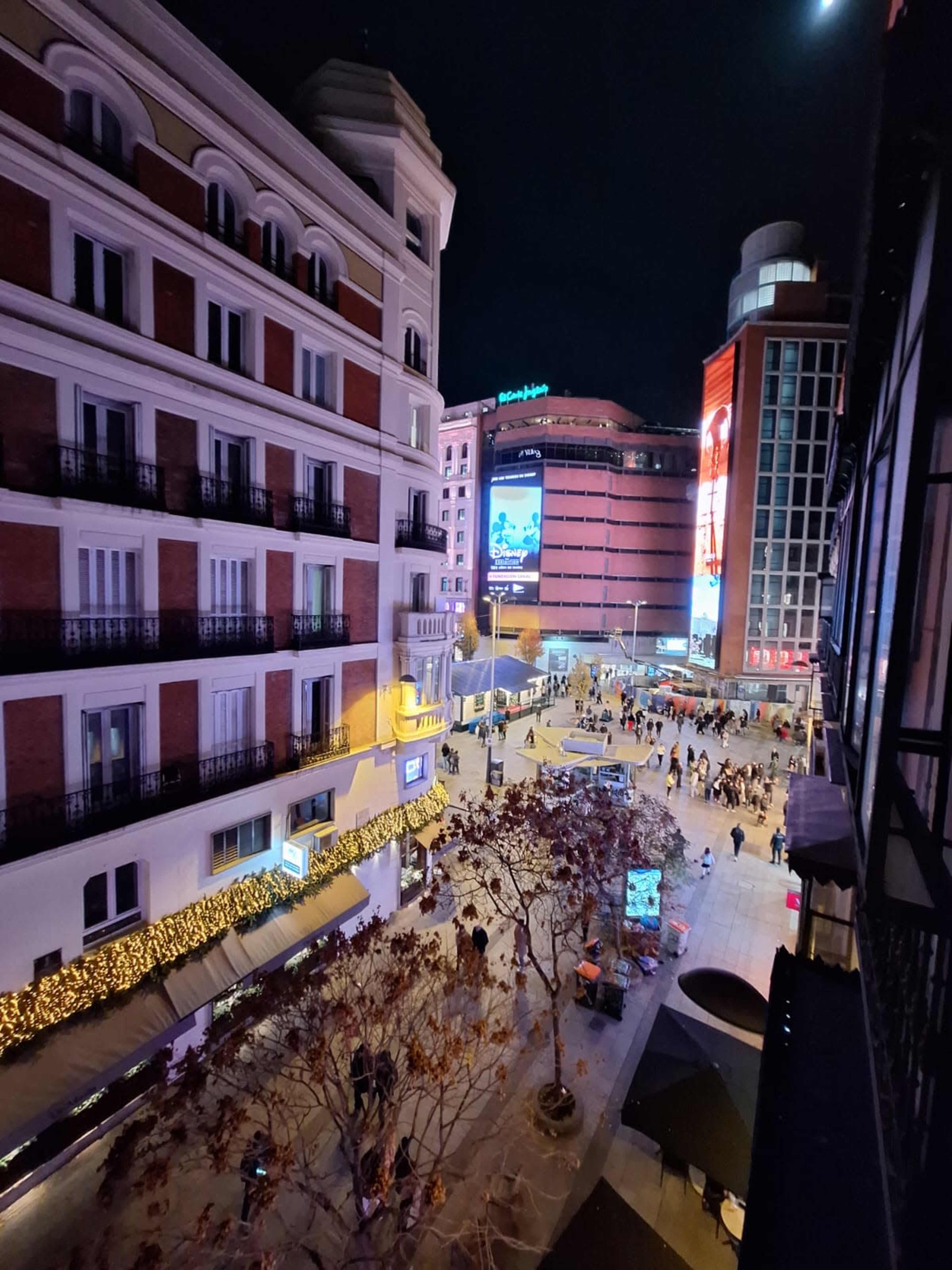 The image shows a bustling urban scene at night, with people walking on a plaza surrounded by buildings and illuminated advertisements.