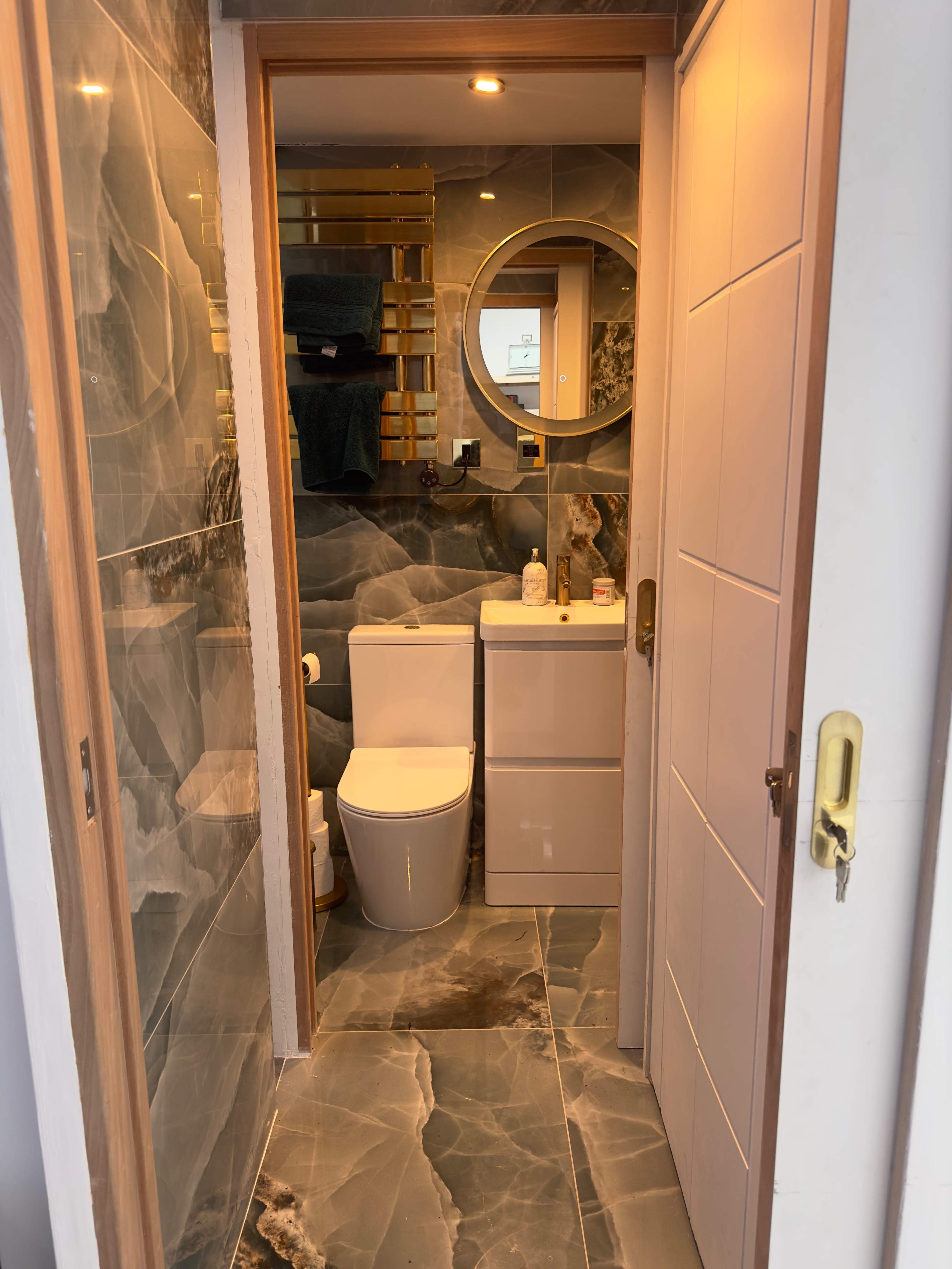 A narrow bathroom with a circular mirror, modern fixtures, and marble-patterned walls and floor tiles.