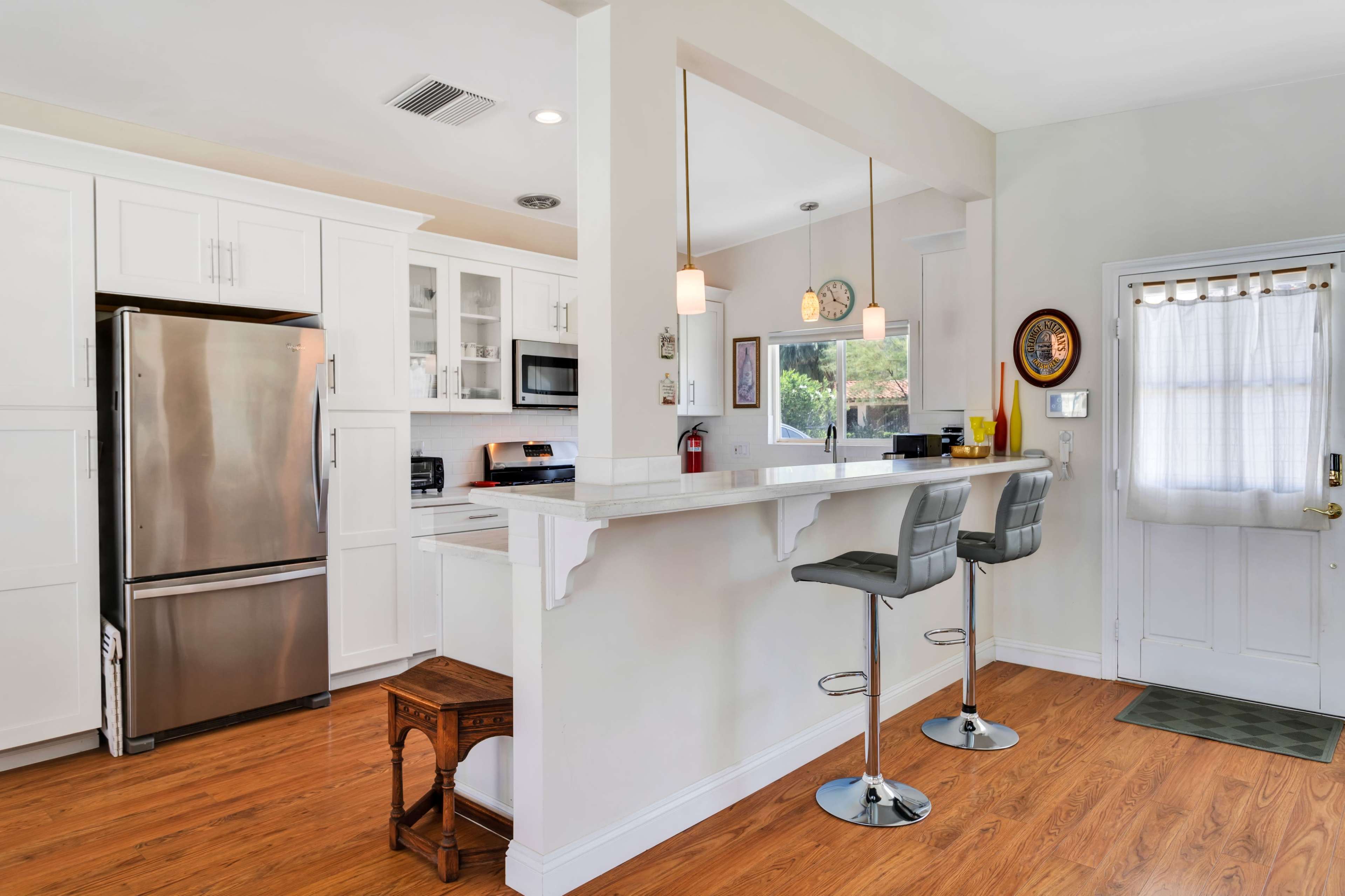 A modern kitchen with a stainless steel refrigerator, white cabinetry, and a bar area with two stools.