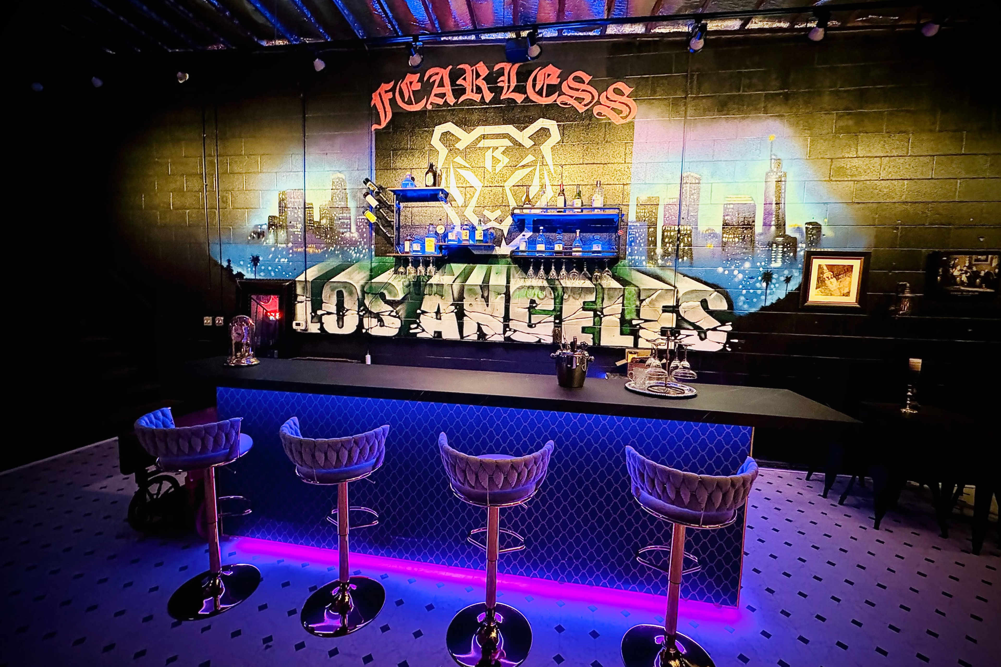 A modern bar area with a mural featuring a bear and "LOS ANGELES," illuminated by blue and purple lighting against a black wall.