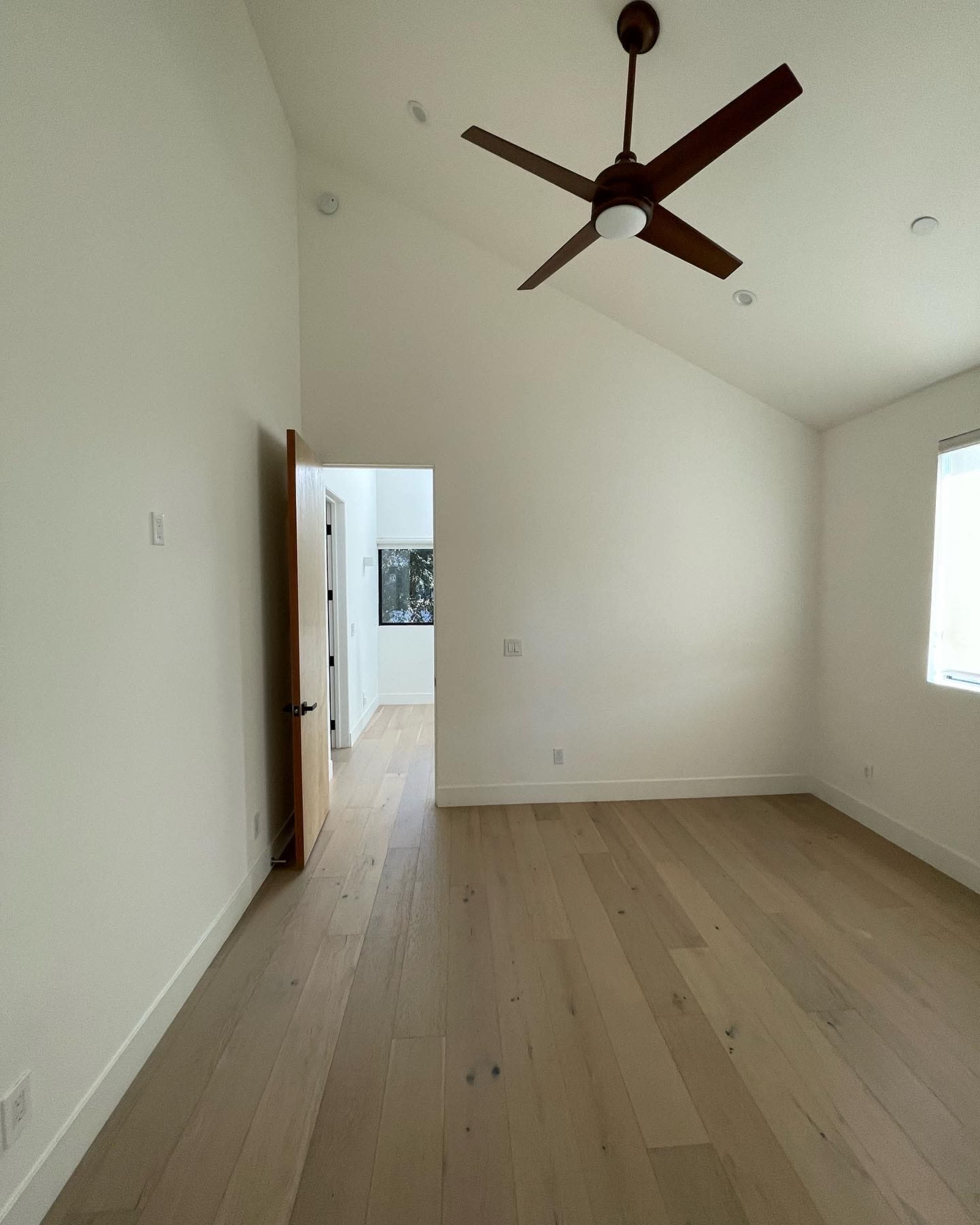 A spacious room with a wooden floor, a ceiling fan, and a doorway leading to another room in the background.