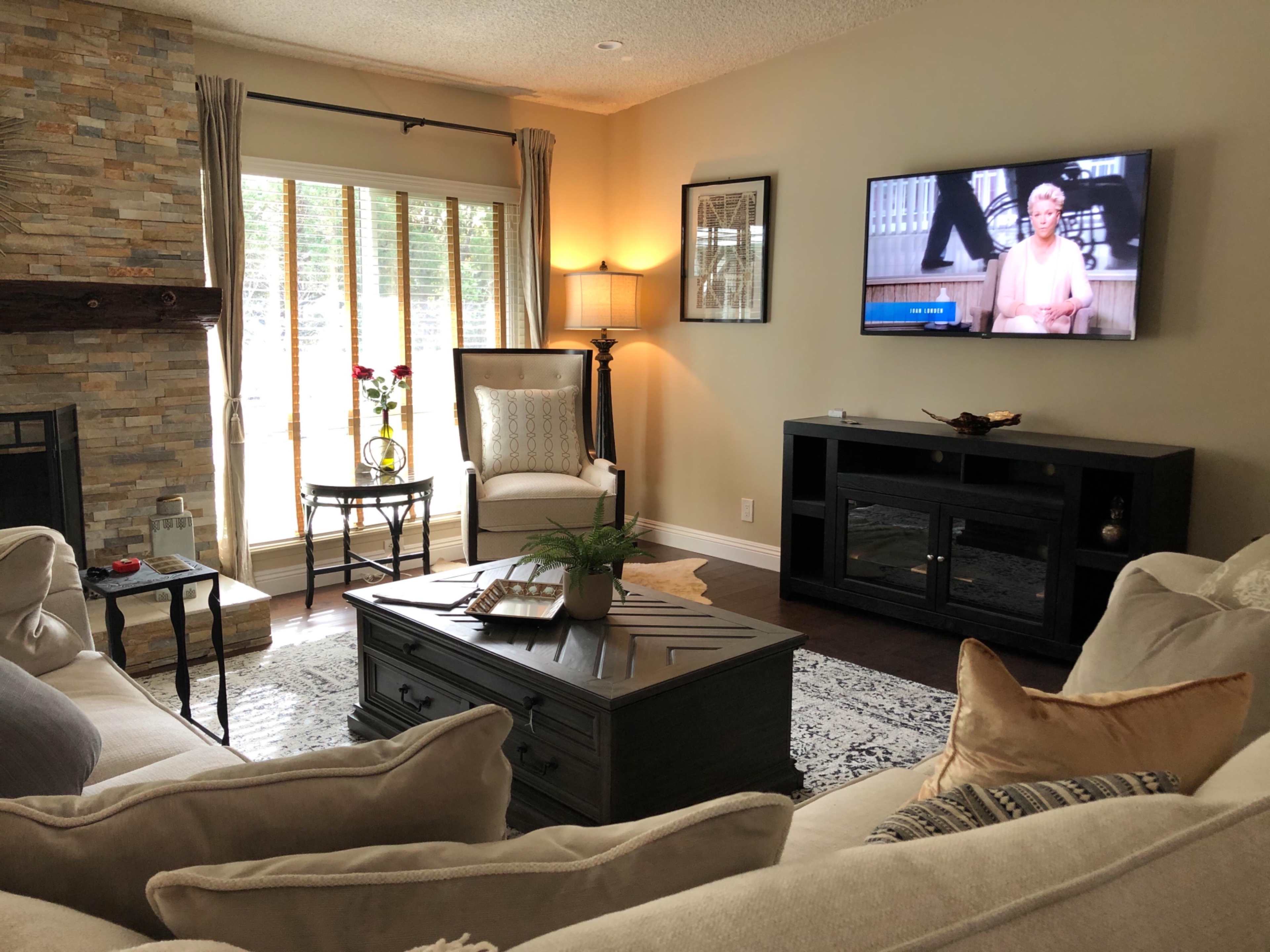 A cozy living room features a large sofa, a coffee table, and a television mounted on the wall, with natural light streaming in through the window.