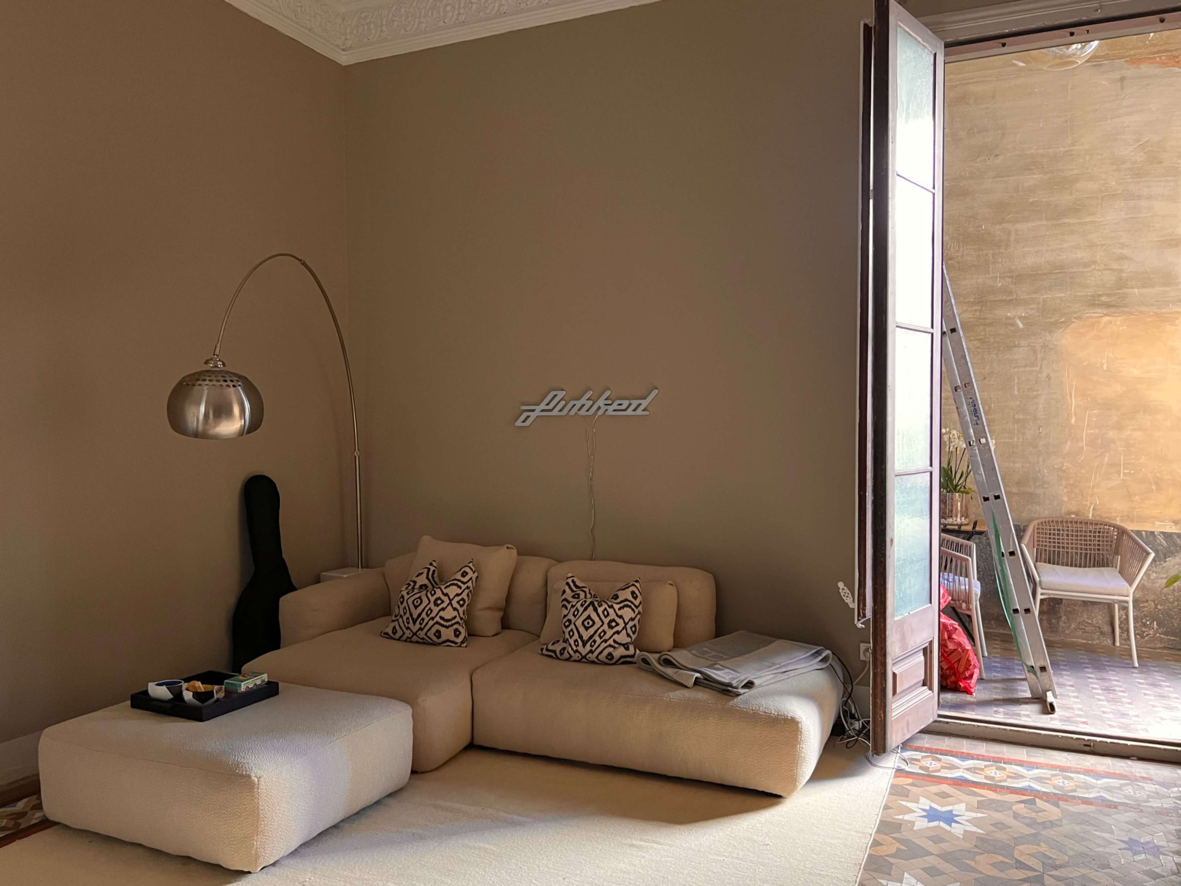 A modern living room features a light-colored sofa, decorative pillows, and an open door leading to an outdoor space with a ladder and chairs.