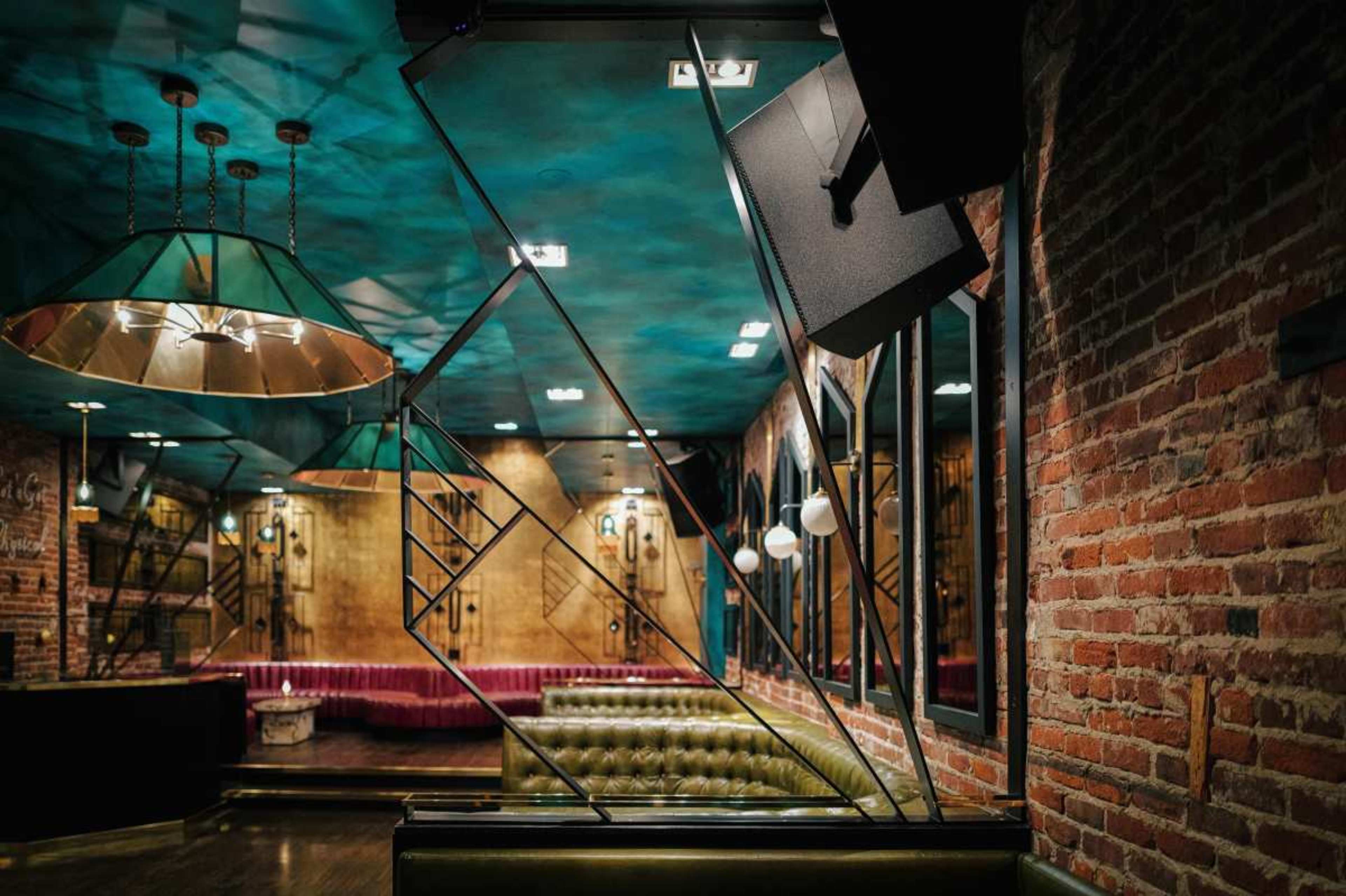 The image shows an interior of a modern bar featuring a green leather couch, exposed brick walls, geometric light fixtures, and a stairway in the background.
