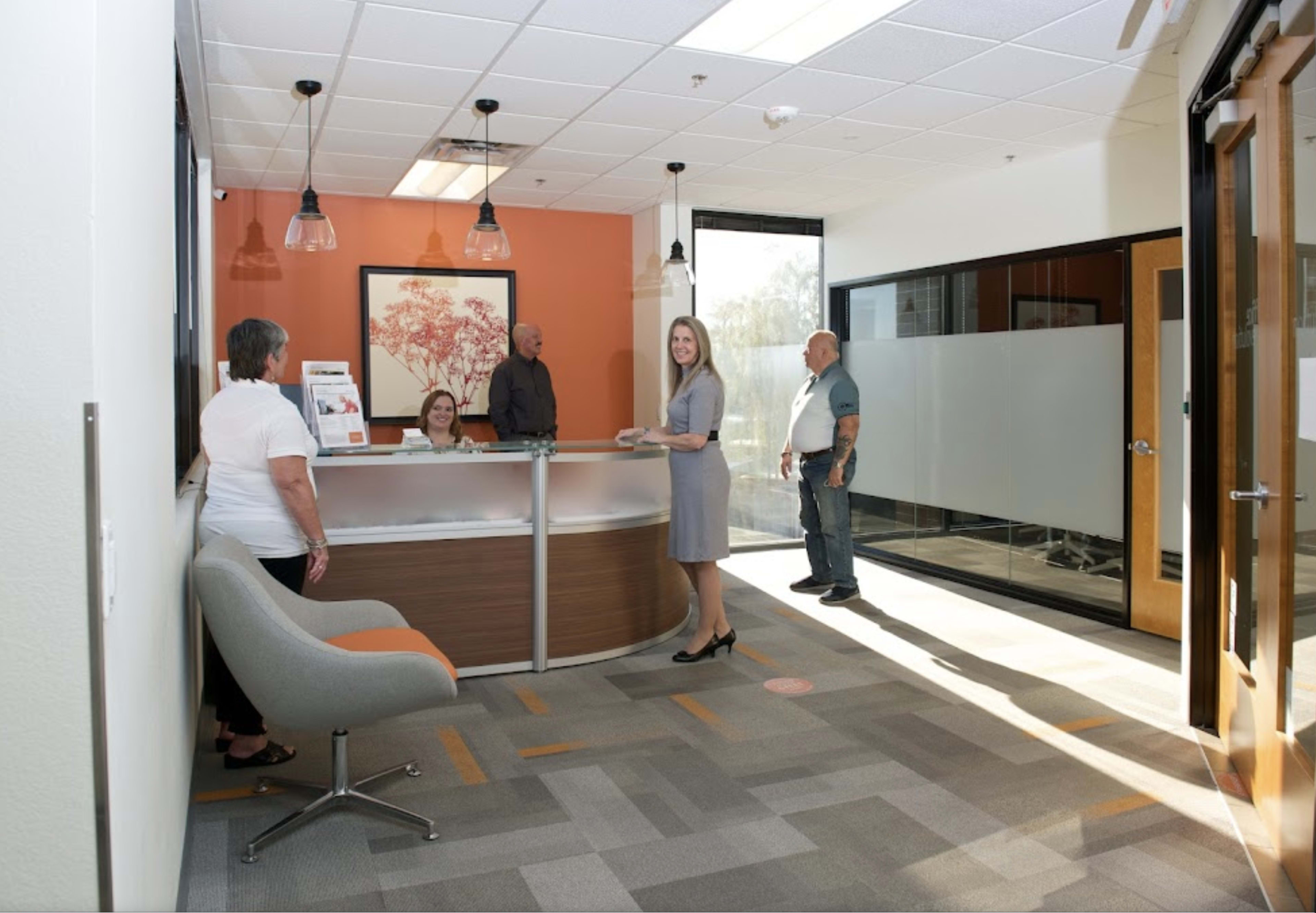 The image shows a reception area with two women and two men interacting, while a woman in a gray dress stands at the desk in front of a modern reception counter.