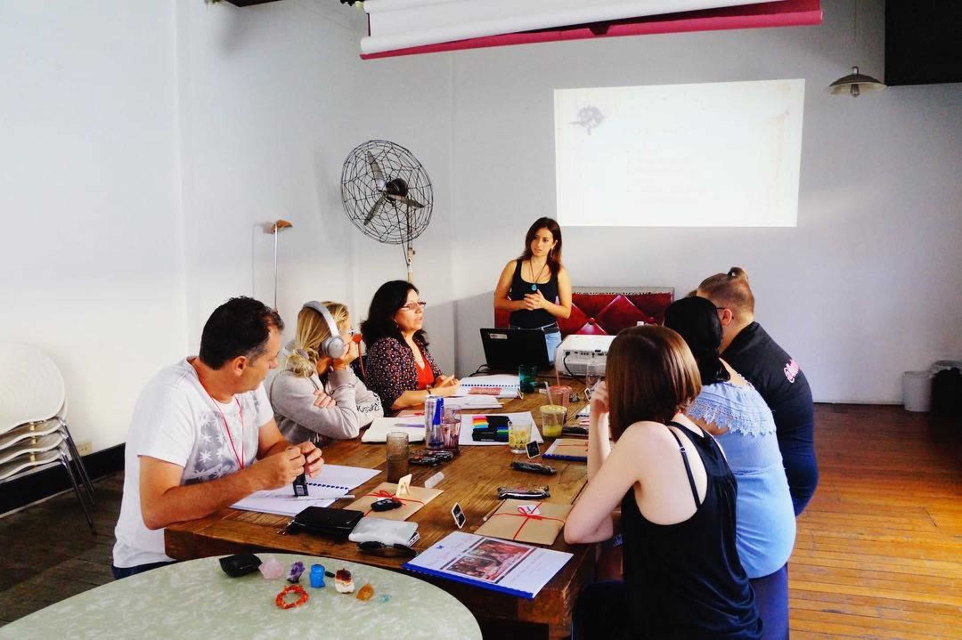 A group of people sits around a table in a well-lit room, while a presenter stands near a projector displaying a presentation on the wall.