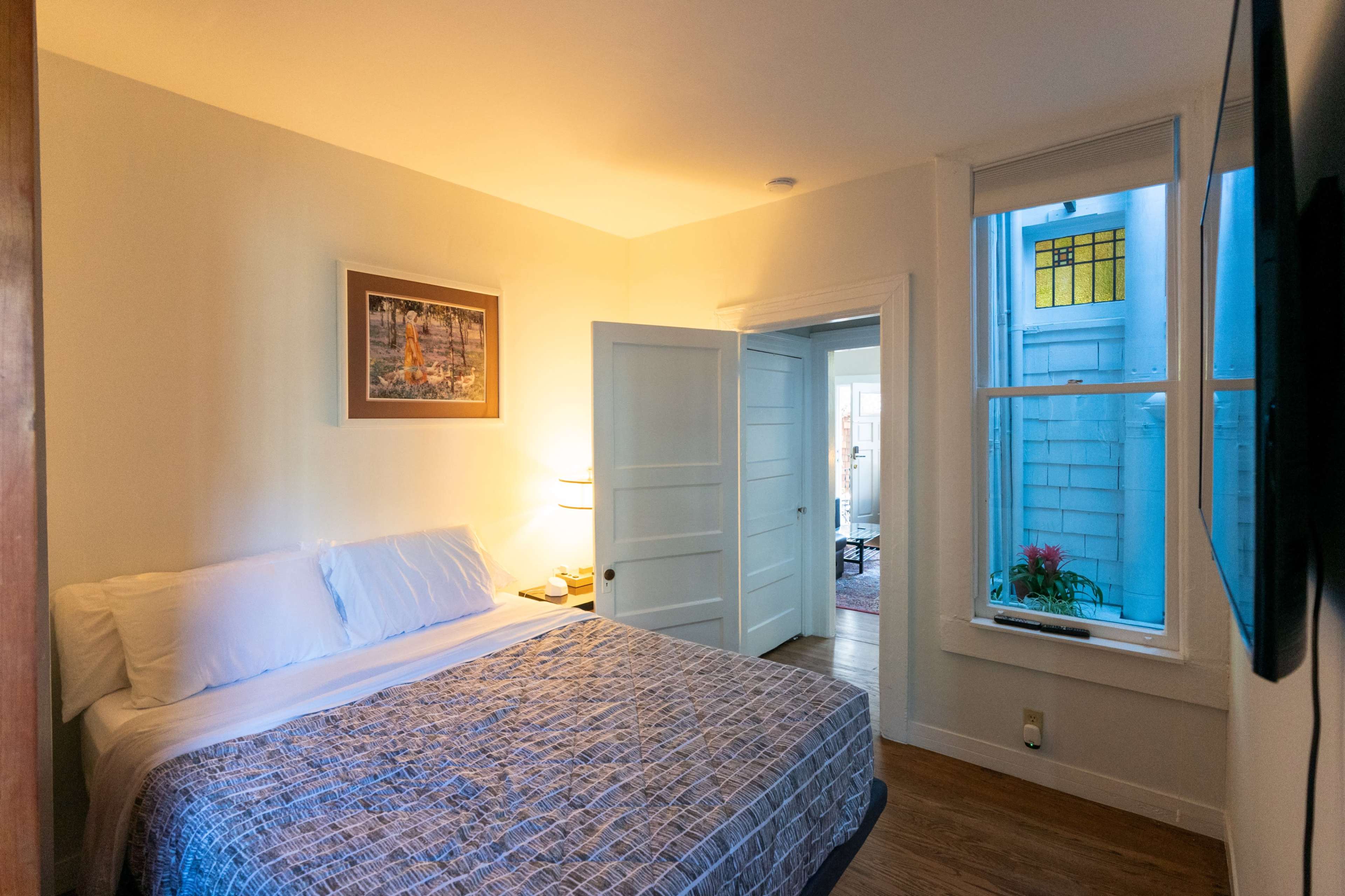 The image shows a small bedroom featuring a bed with white linens, a bedside lamp, and a colorful piece of stained glass visible through a nearby window.