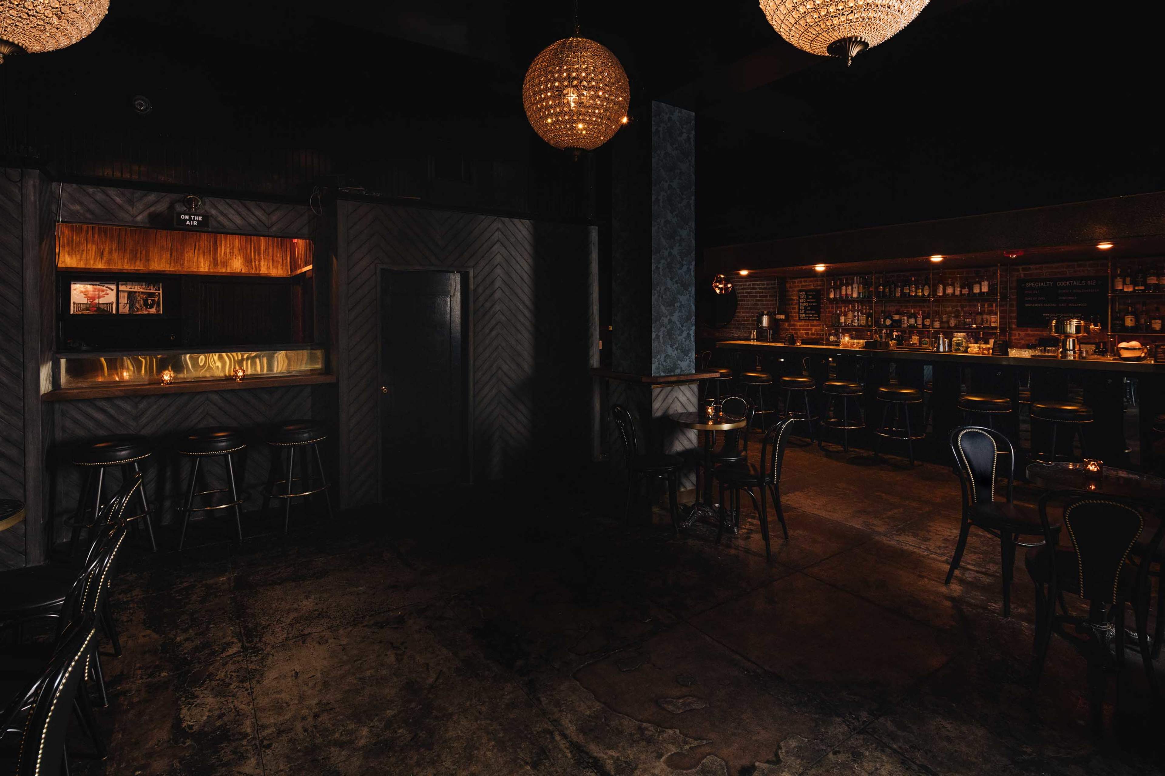 A dimly lit bar features dark furniture and a long counter illuminated by pendant lights.