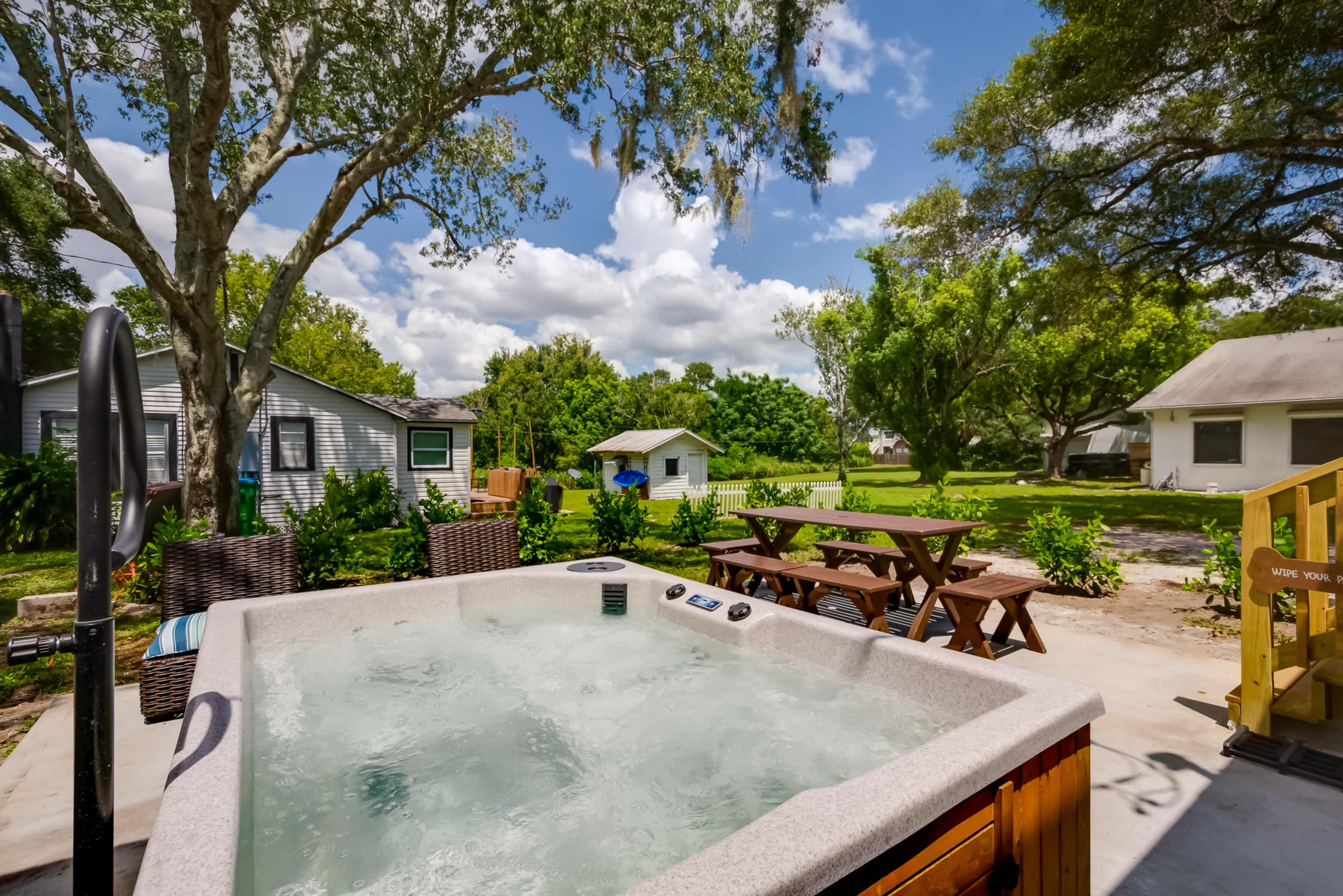 A hot tub is positioned on a patio overlooking a green yard with picnic tables and nearby houses.
