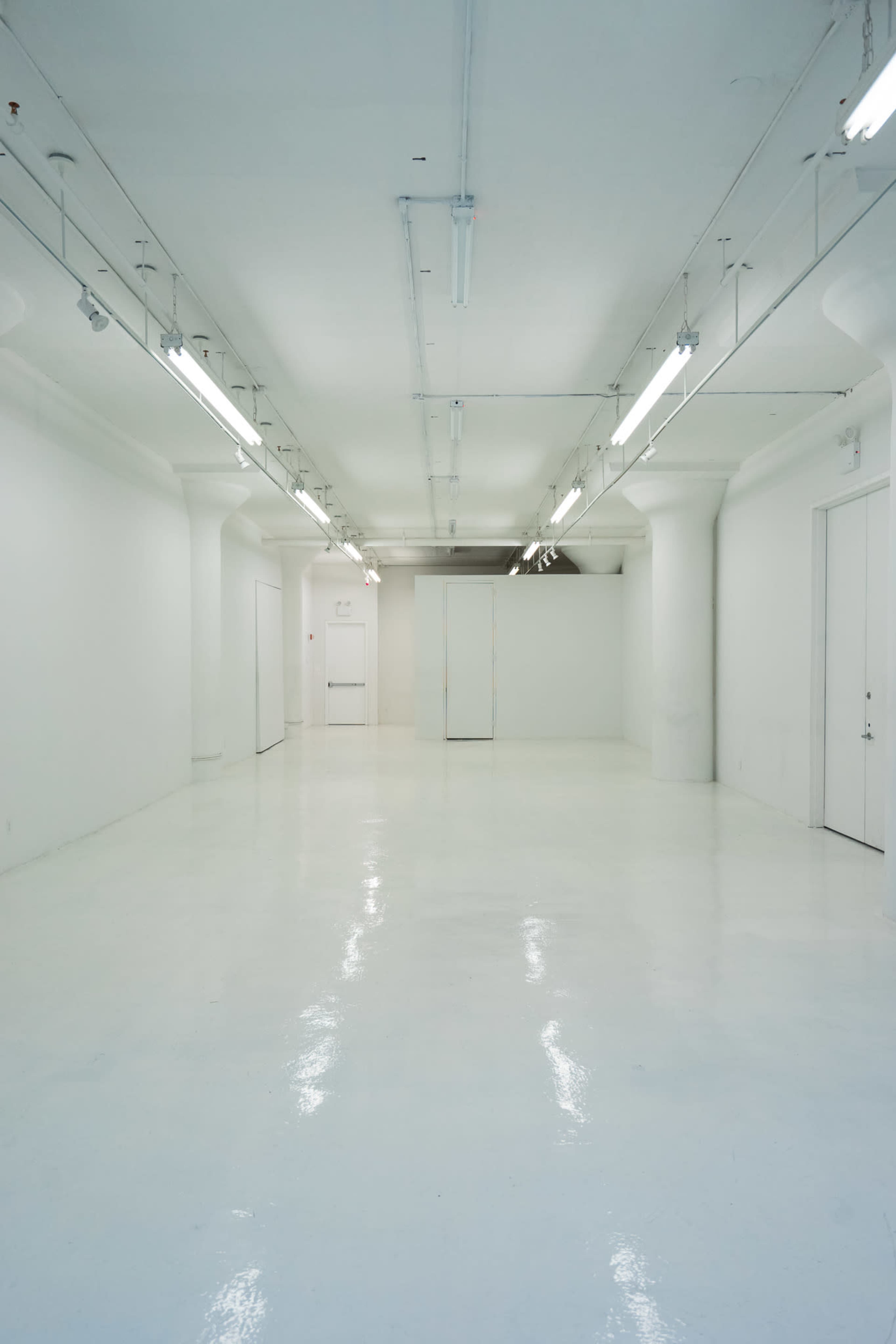 The image shows a large, empty white room with polished floors, bright overhead lights, and doors at one end.