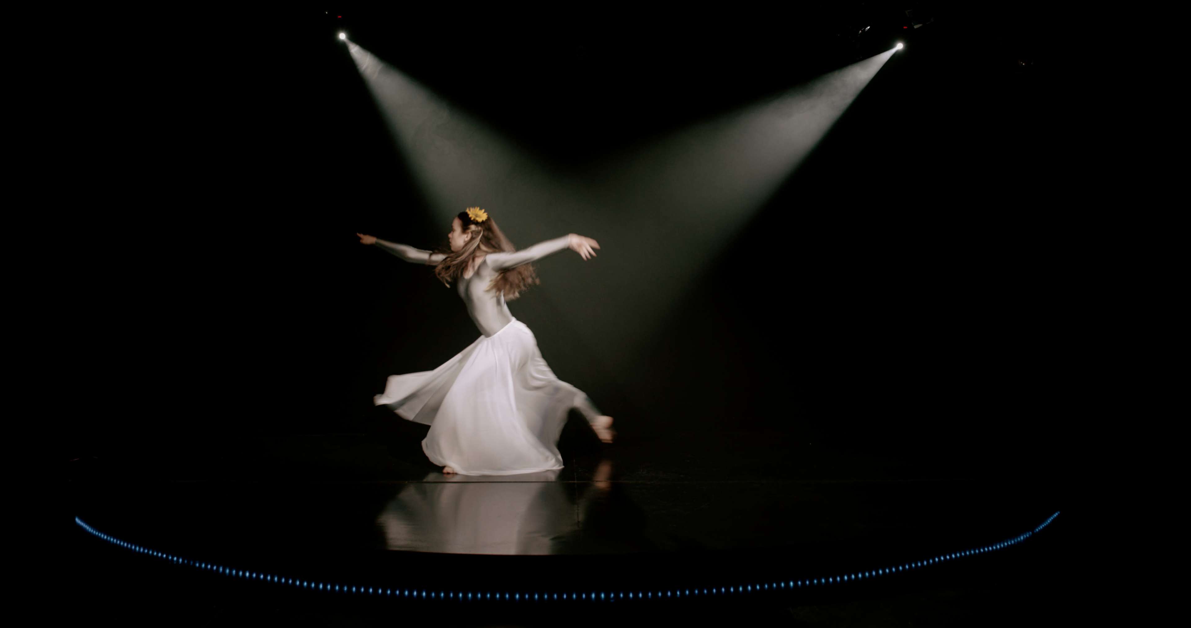 A dancer in a flowing white dress spins gracefully on a dimly lit stage, illuminated by pointed beams of light.