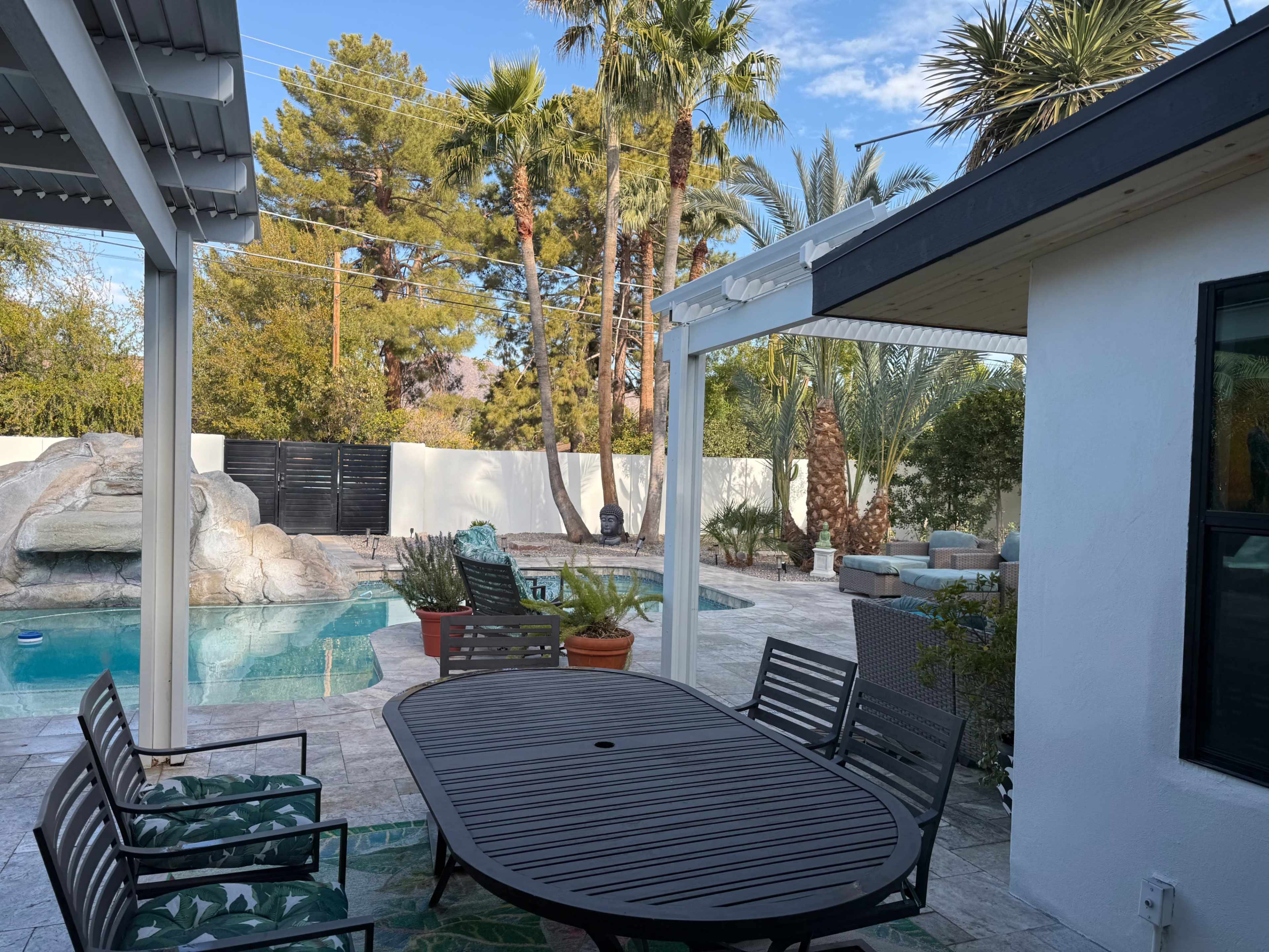The image depicts a backyard with a swimming pool, palm trees, and a stone feature, complemented by a dining table and chairs under a covered patio area.