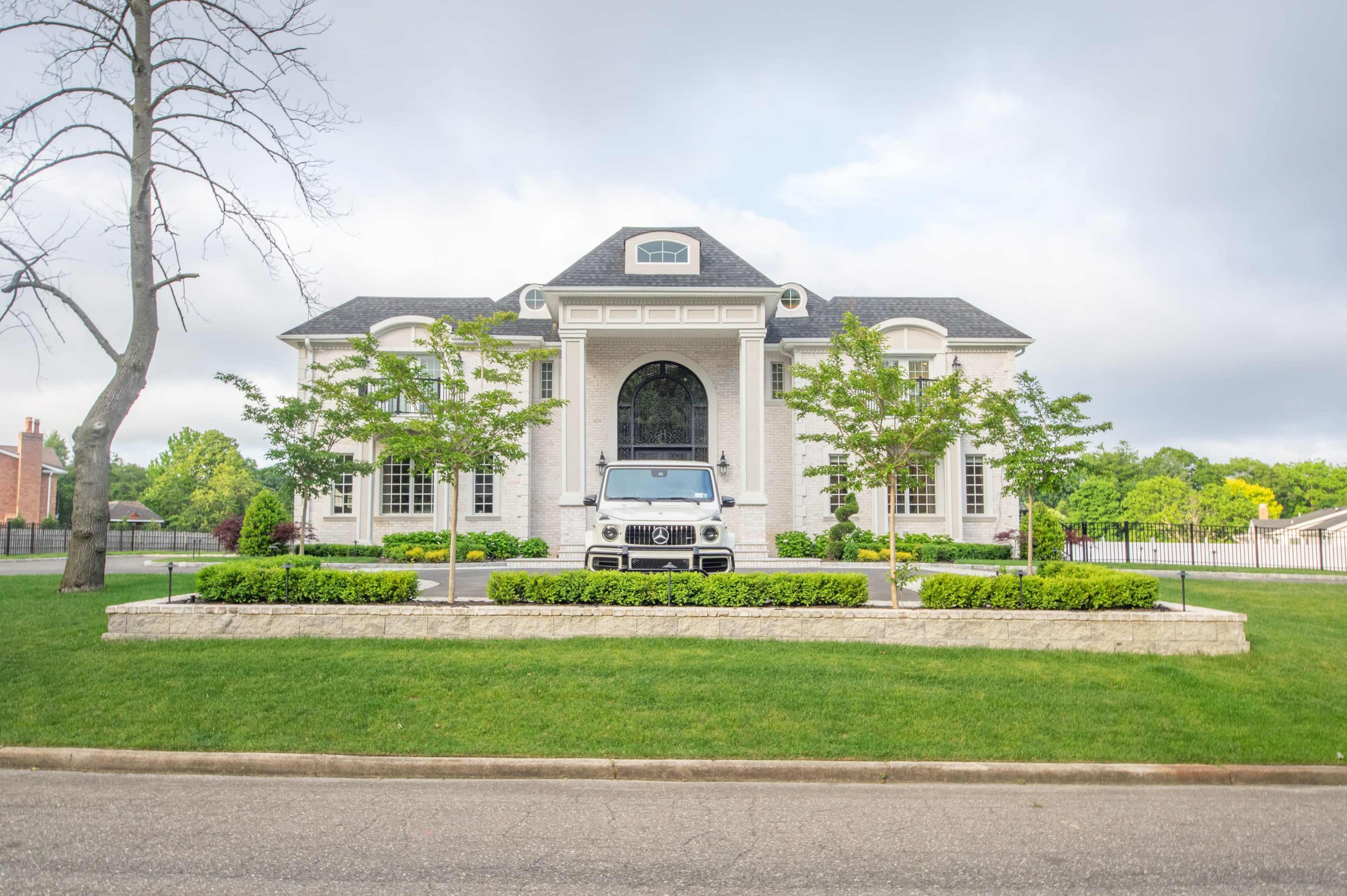 A large, white brick mansion with a front driveway and a parked vehicle is framed by well-maintained landscaping.