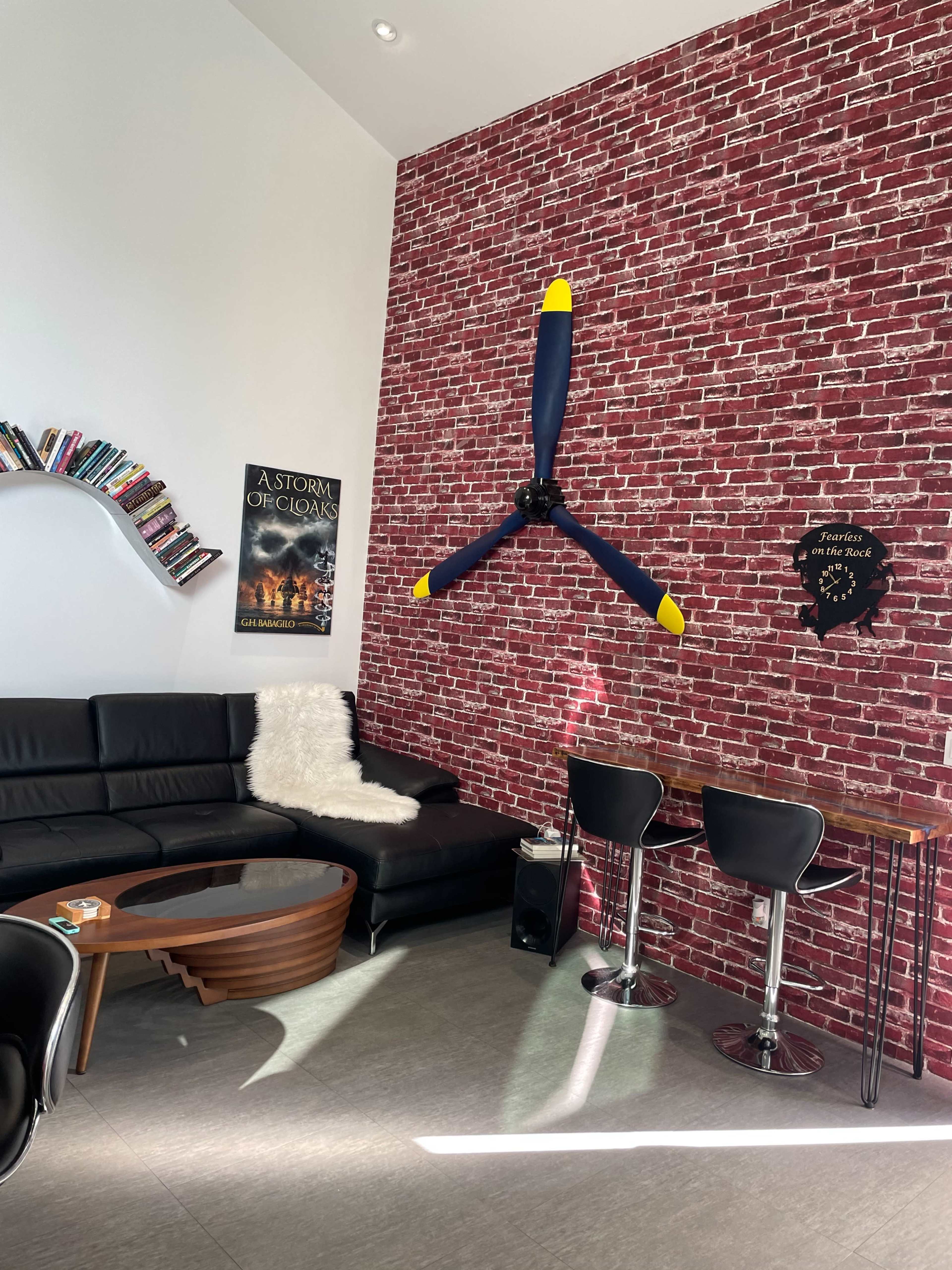 The image shows a modern living area featuring a black leather sofa, a wooden coffee table, a wall with a brick pattern, and a large propeller mounted as a decorative piece.