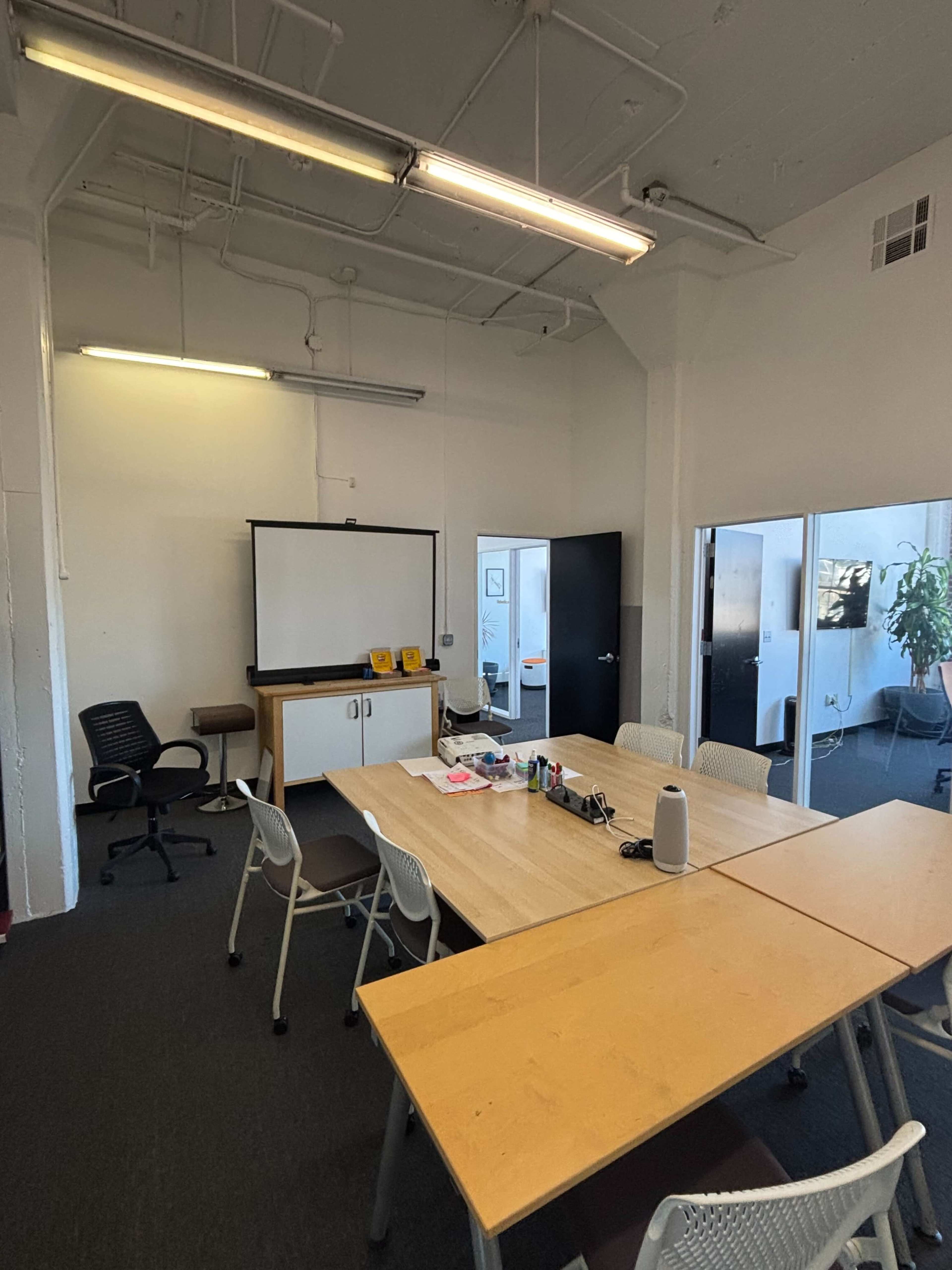 A modern conference room features a large wooden table surrounded by chairs, a wall-mounted screen, and a doorway leading to another room.