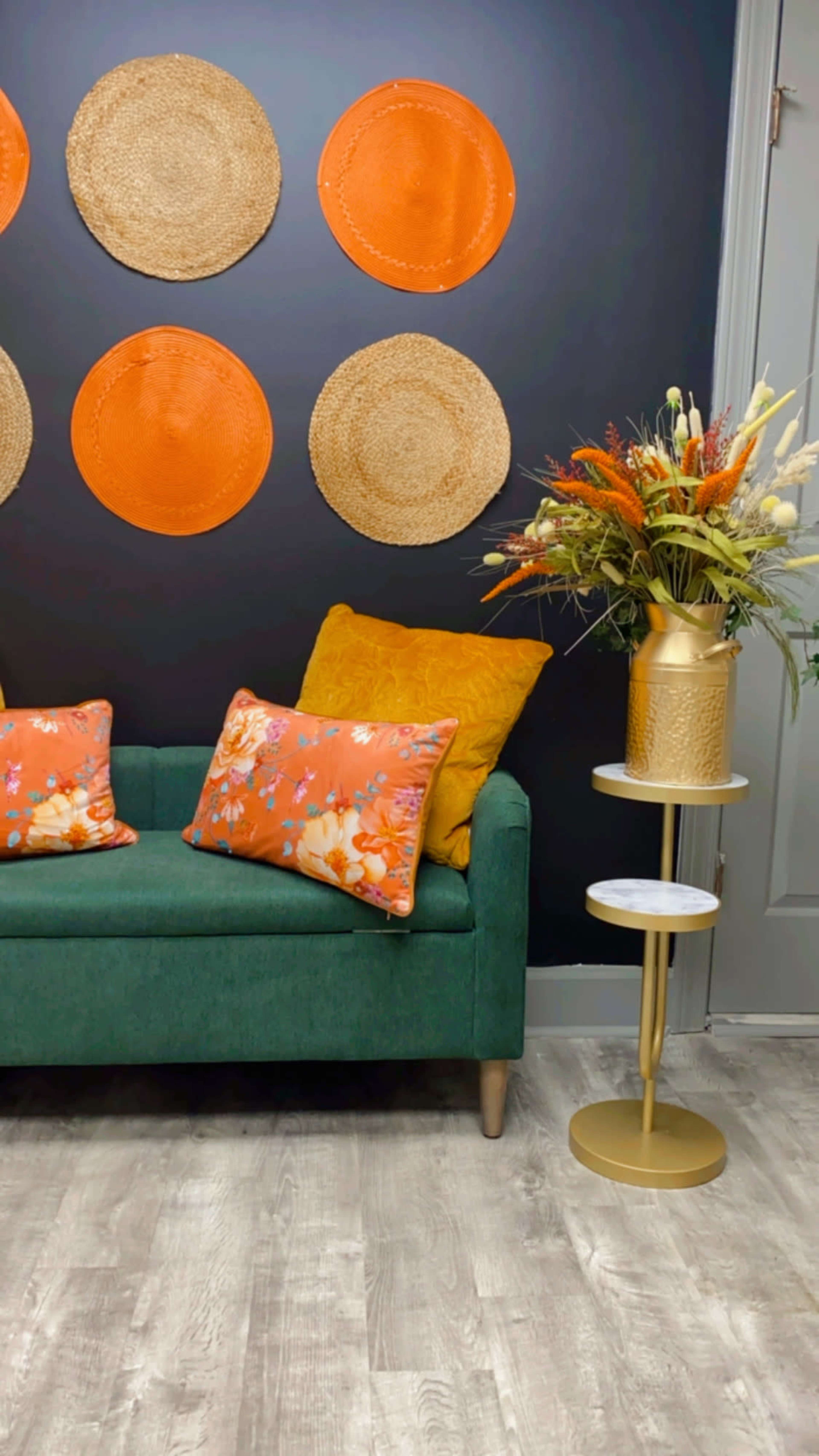 A green sofa with patterned orange and yellow cushions is positioned against a dark wall adorned with woven circular wall art, next to a gold side table topped with a floral arrangement.
