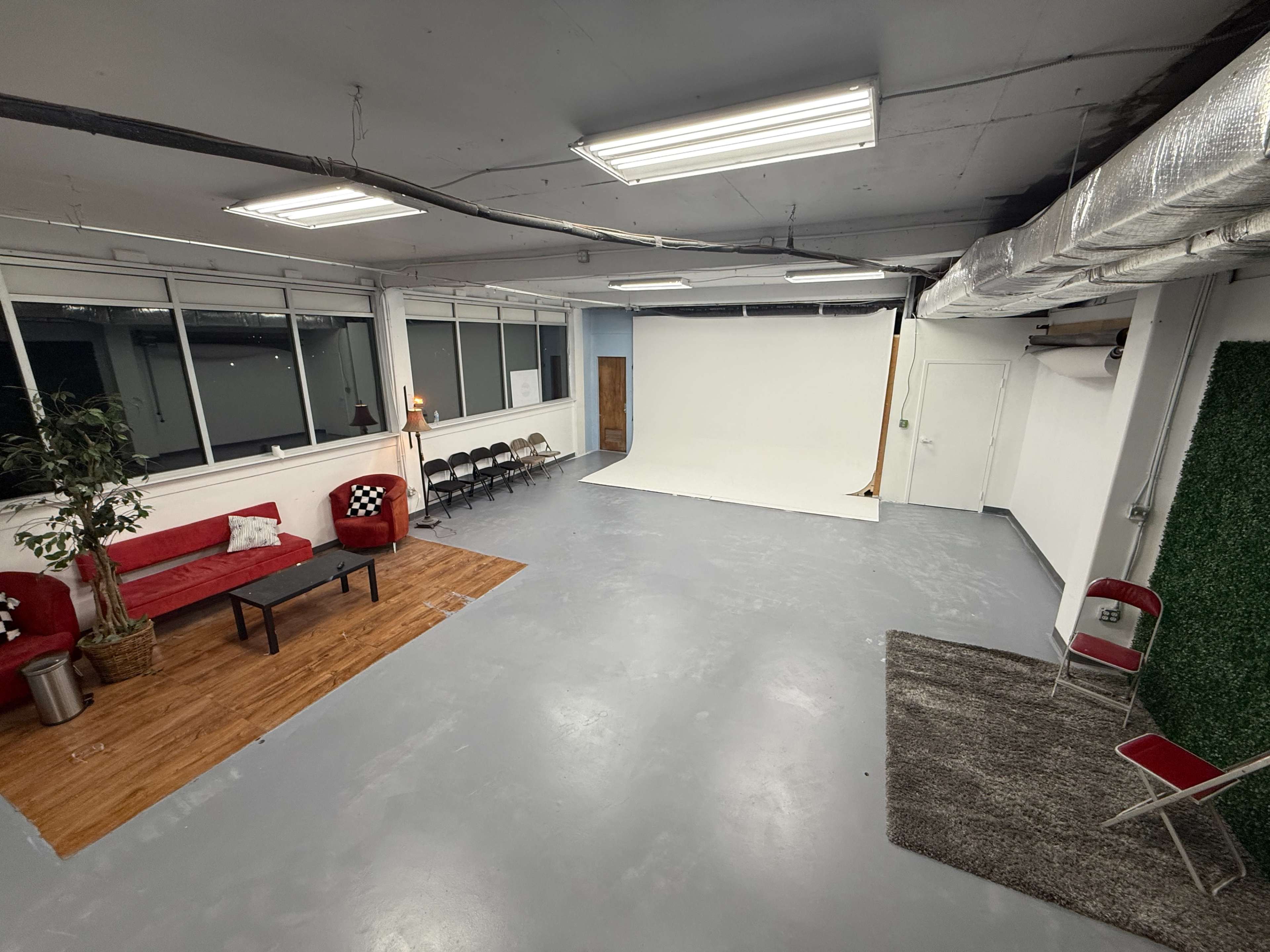 The image shows a spacious studio with a white backdrop, wooden flooring, several chairs arranged in rows, and a small seating area with a red couch and a plant.