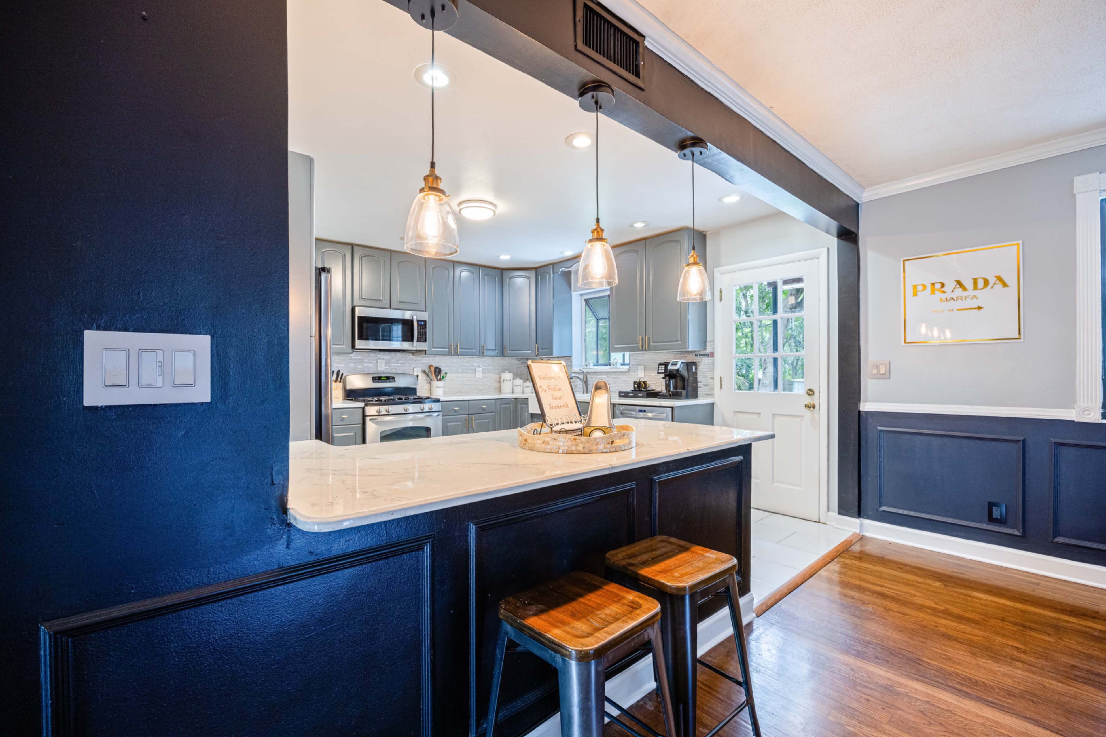 A modern kitchen features gray cabinets, stainless steel appliances, and a bar area with three wooden stools illuminated by pendant lights.