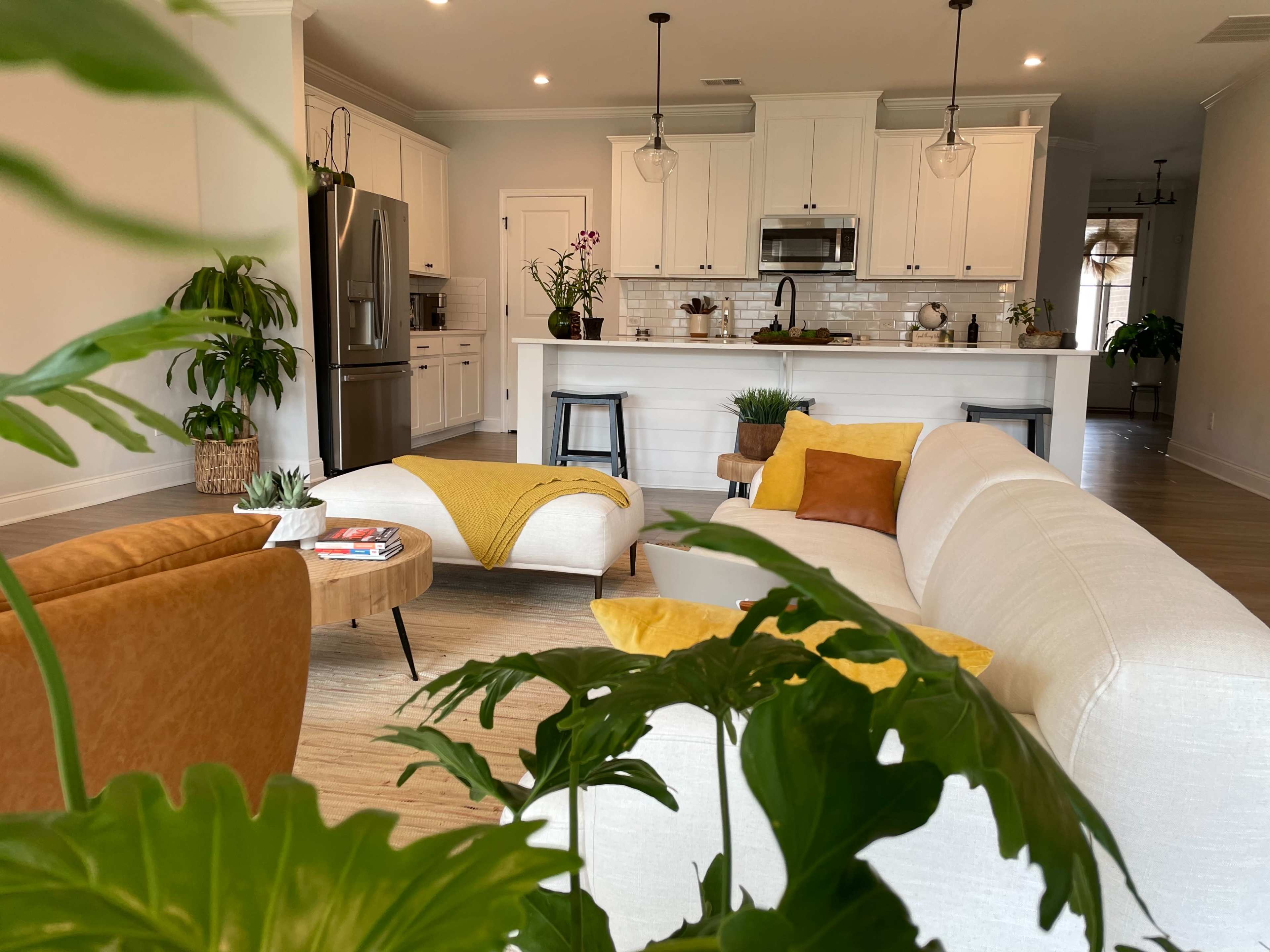 A living room features a beige sofa, orange armchair, and a round wooden coffee table, with a view of a kitchen in the background.