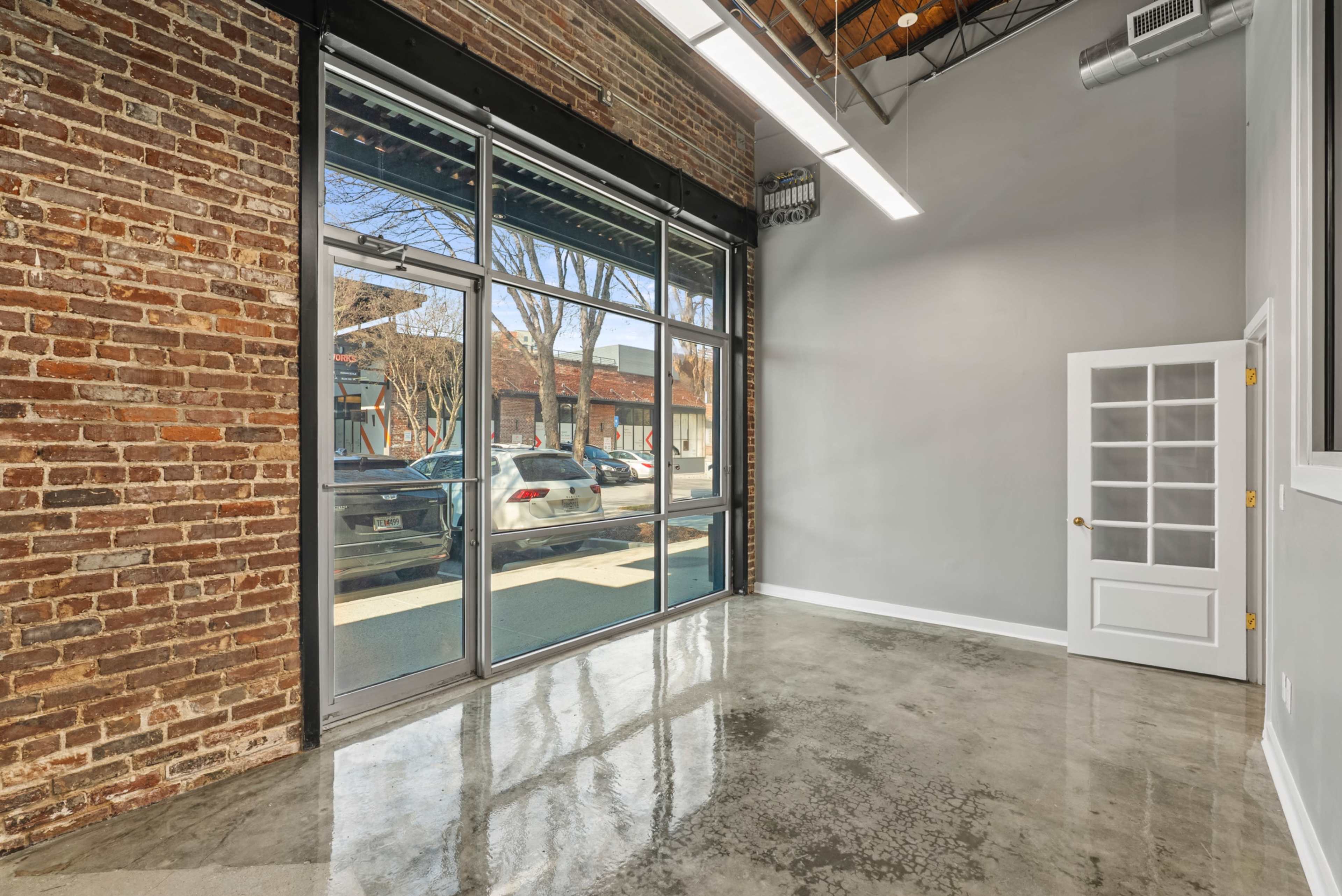 The image shows an empty, modern interior space with exposed brick walls, large glass windows, and a polished concrete floor.