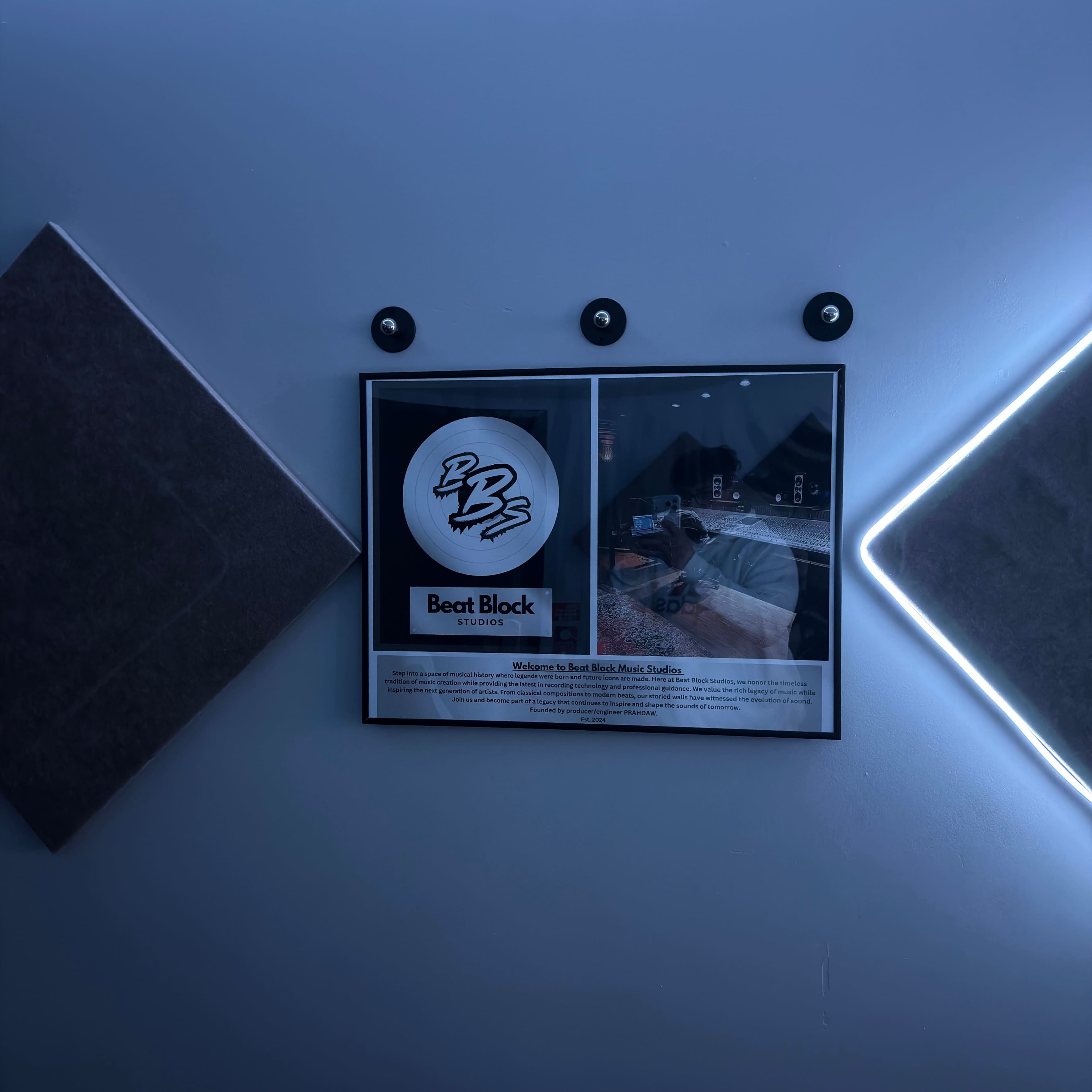 The image shows a framed poster on a wall that promotes "Beat Block Studios," accompanied by three circular wall fixtures and two square acoustic panels.
