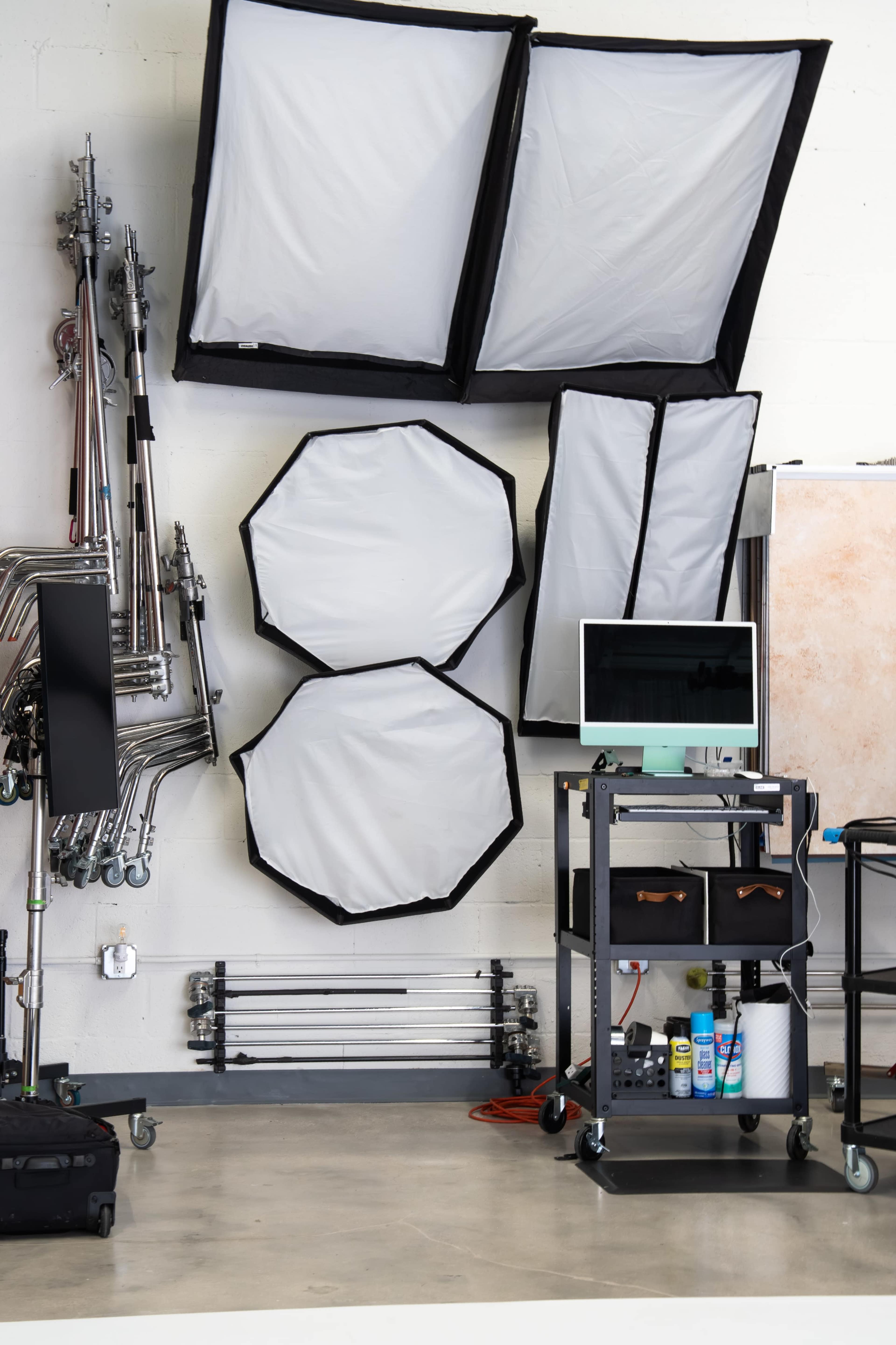 The image shows a studio setup with various lighting equipment, including softboxes, stands, and a cart with supplies, arranged against a wall.