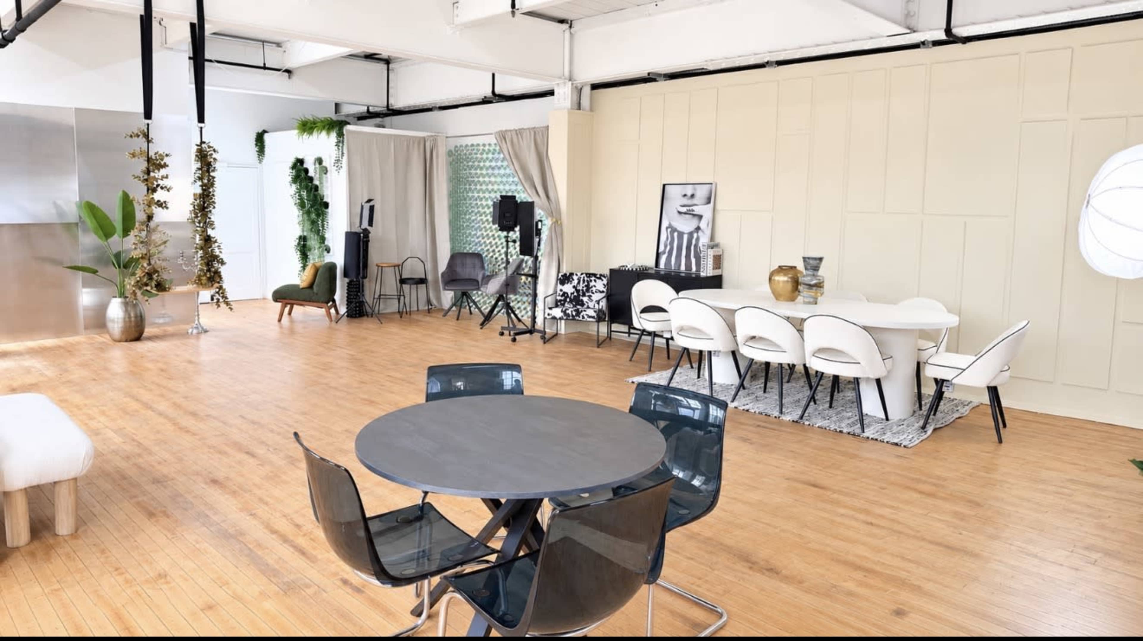 The image shows an open, well-lit interior space featuring a dining area with a large white table, chairs, and a circular table with black chairs, along with various decorative plants and furnishings.