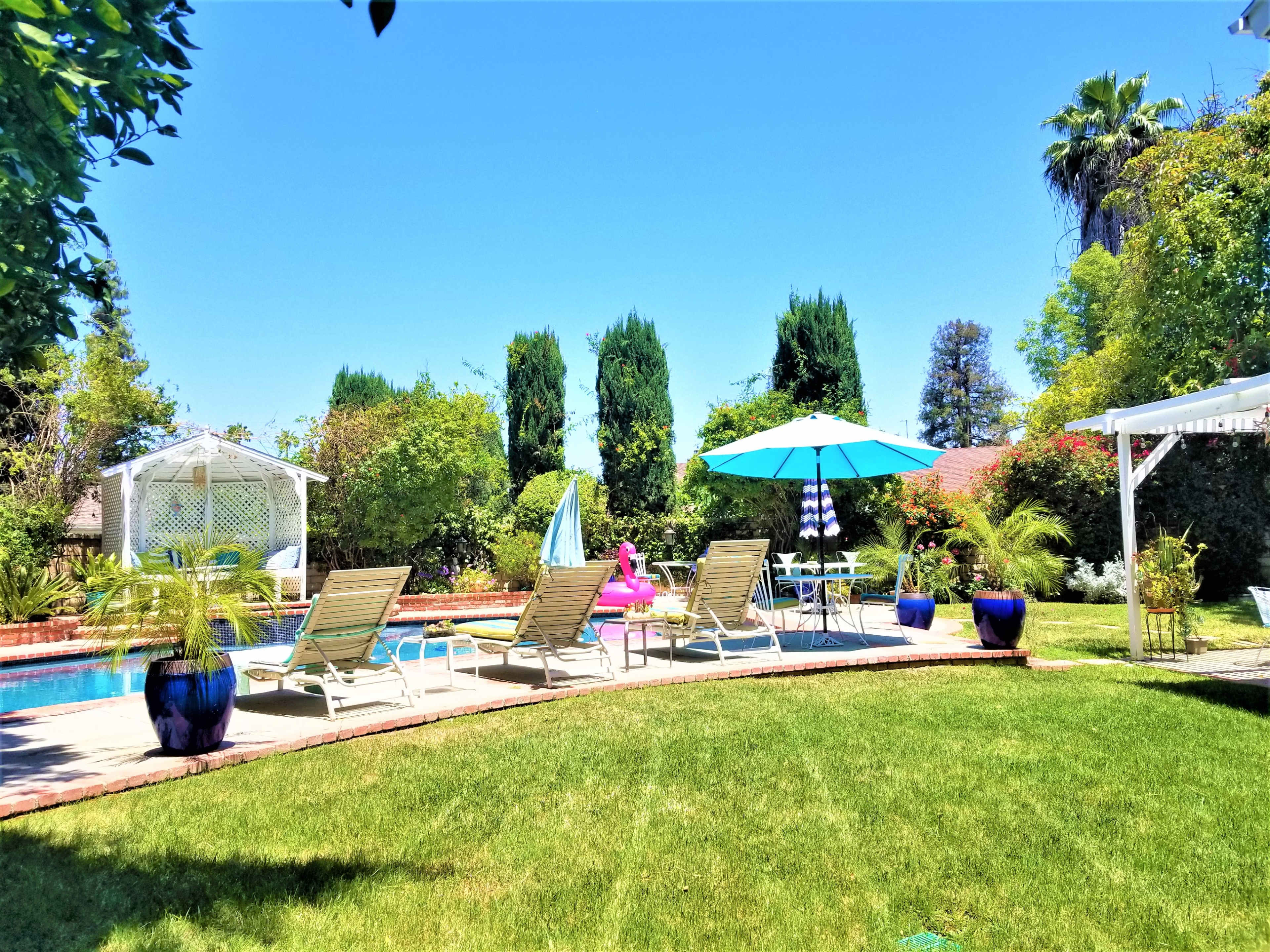 A backyard pool area features lounge chairs, an umbrella, and lush greenery under a clear blue sky.