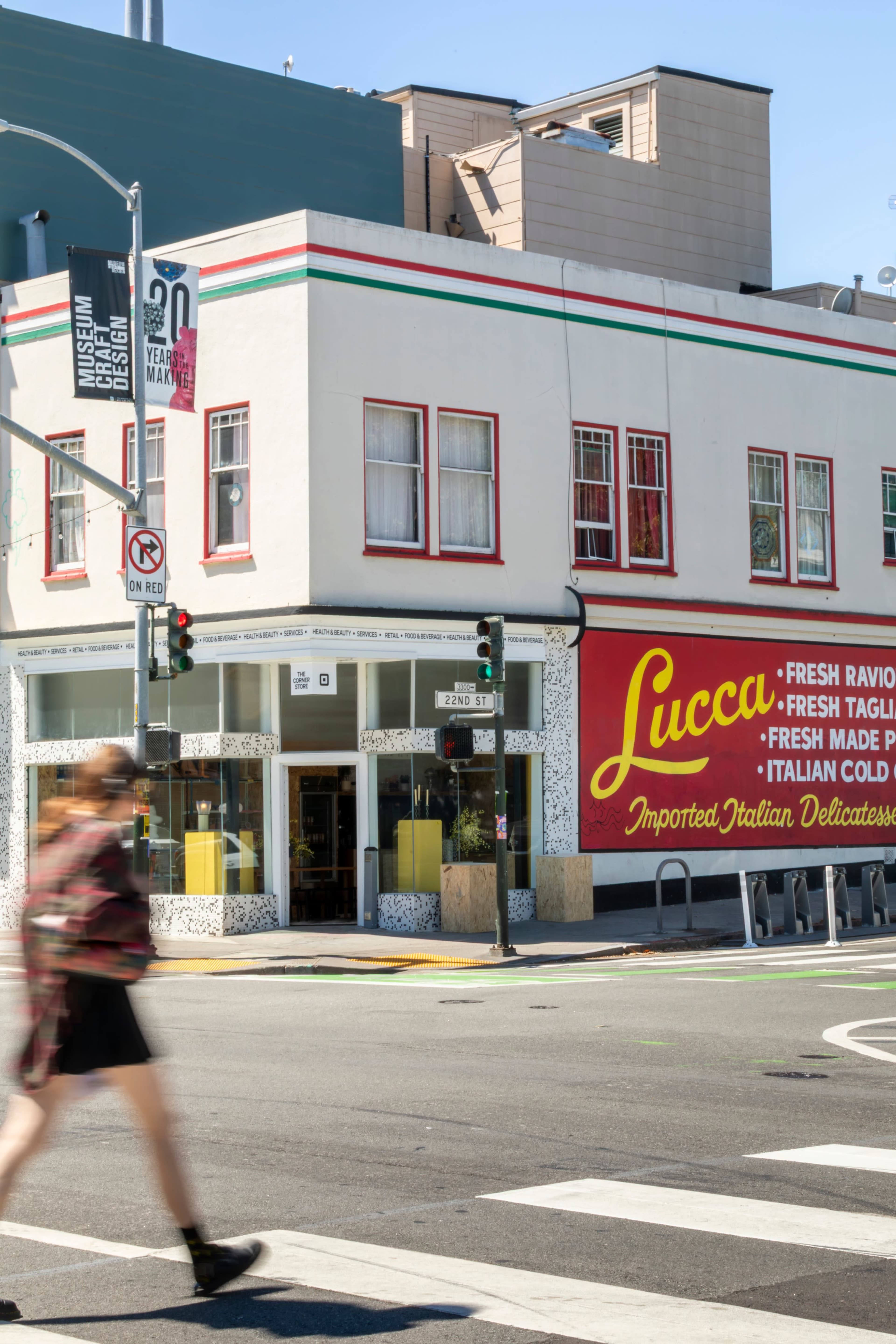 A person walks past a building featuring a prominent sign advertising "Lucca" and its imported Italian delicacies, with a mix of retail signage visible on the façade.