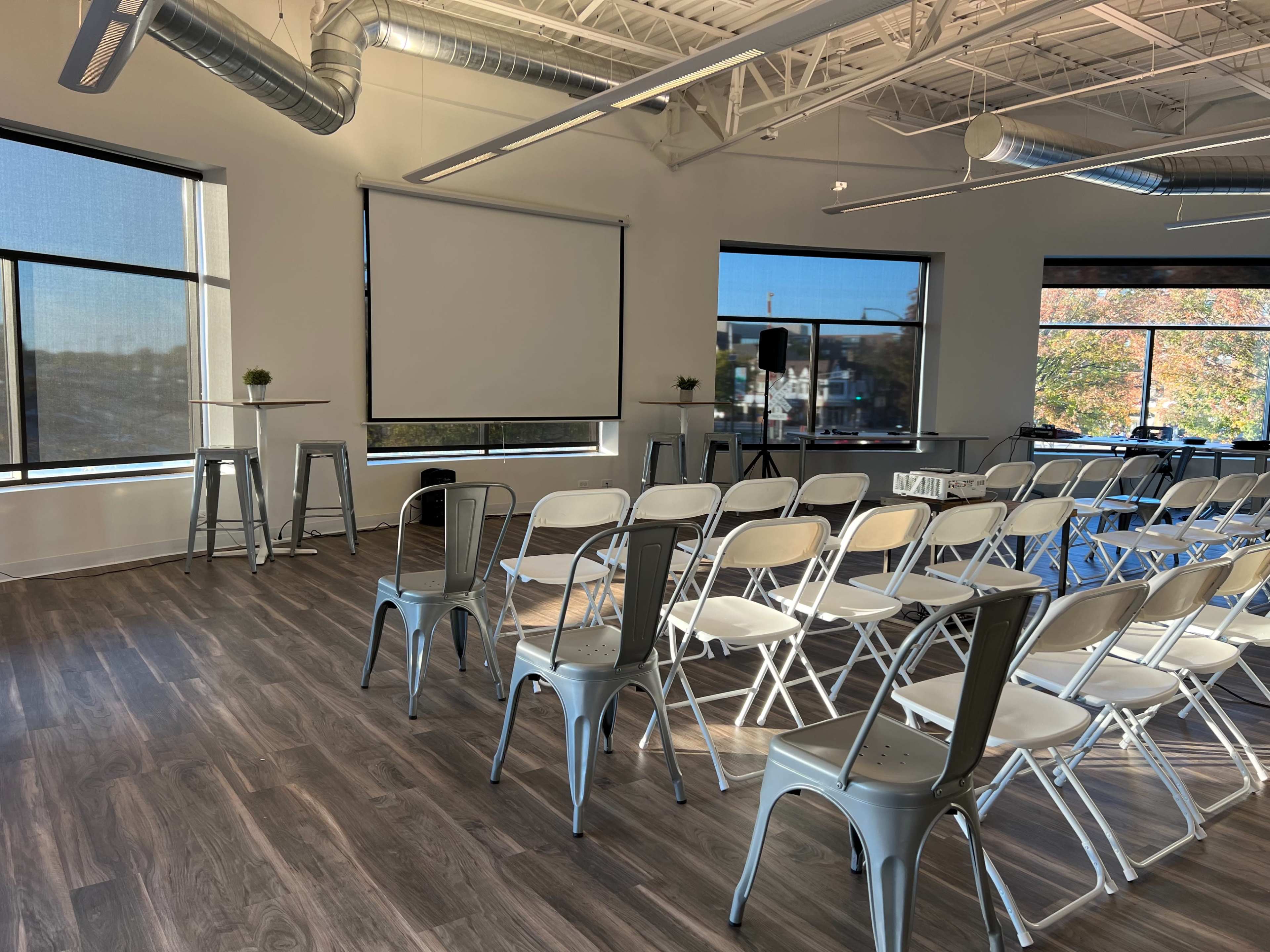 A conference room set up with rows of chairs facing a blank presentation screen, alongside a table on a stage and large windows revealing an outdoor view.