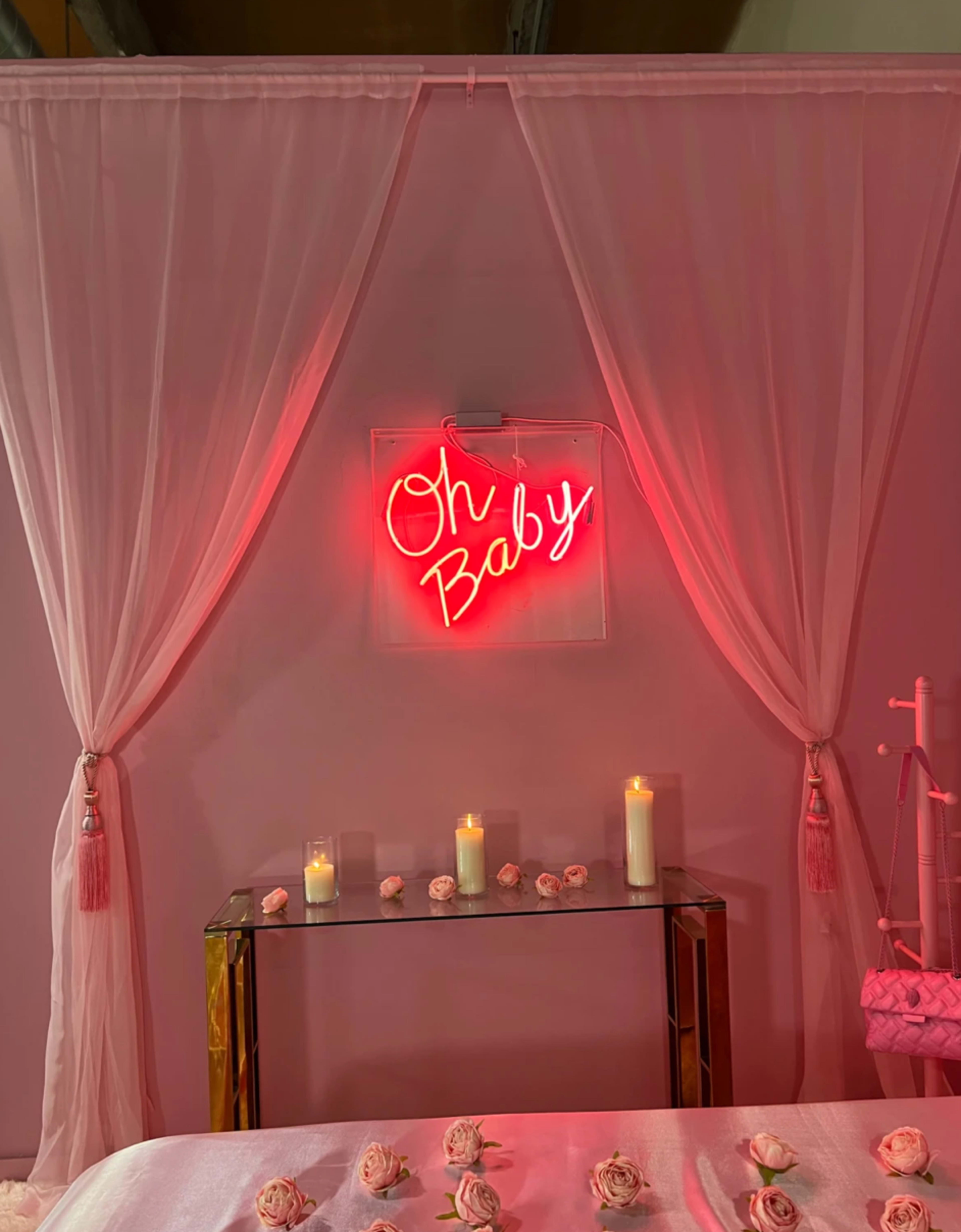 A softly lit room with pink curtains, a neon sign that says "Oh Baby," candles on a glass table, and rose petals scattered on a bed.