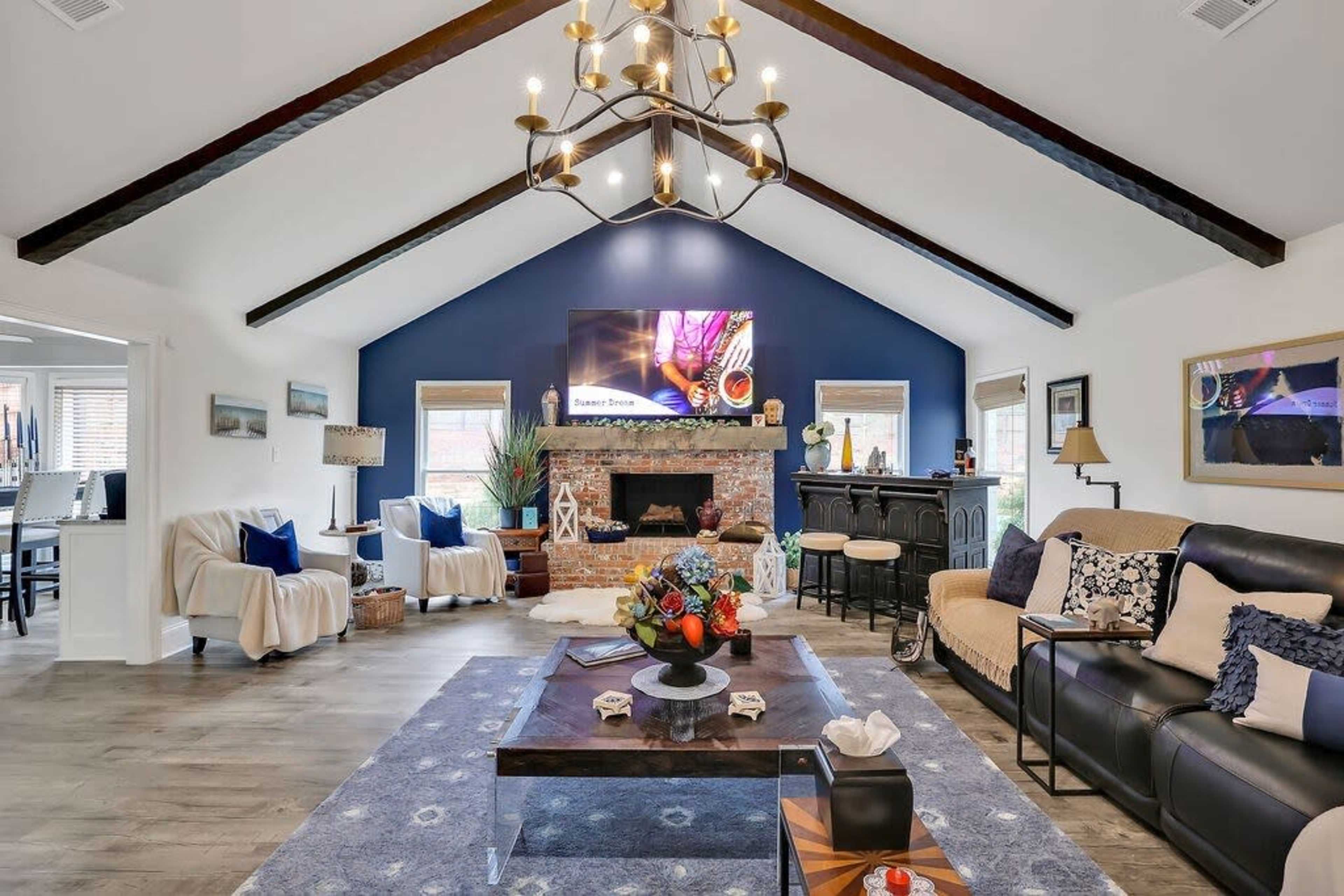 The living room features a vaulted ceiling with exposed beams, a large flat-screen TV mounted on a dark blue wall, and a central coffee table surrounded by sofas and decorative accents.