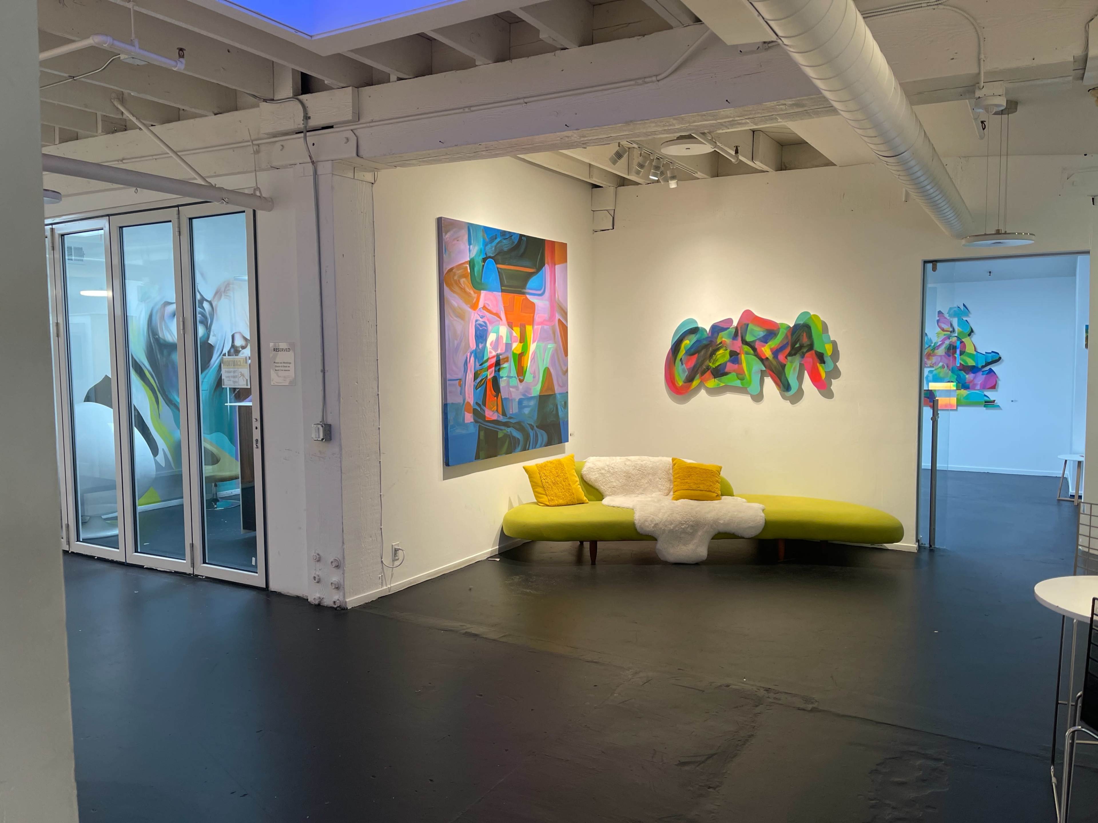 The scene features a brightly colored abstract painting on the wall alongside a vibrant mural, with a yellow-accented green couch positioned in a modern gallery space.