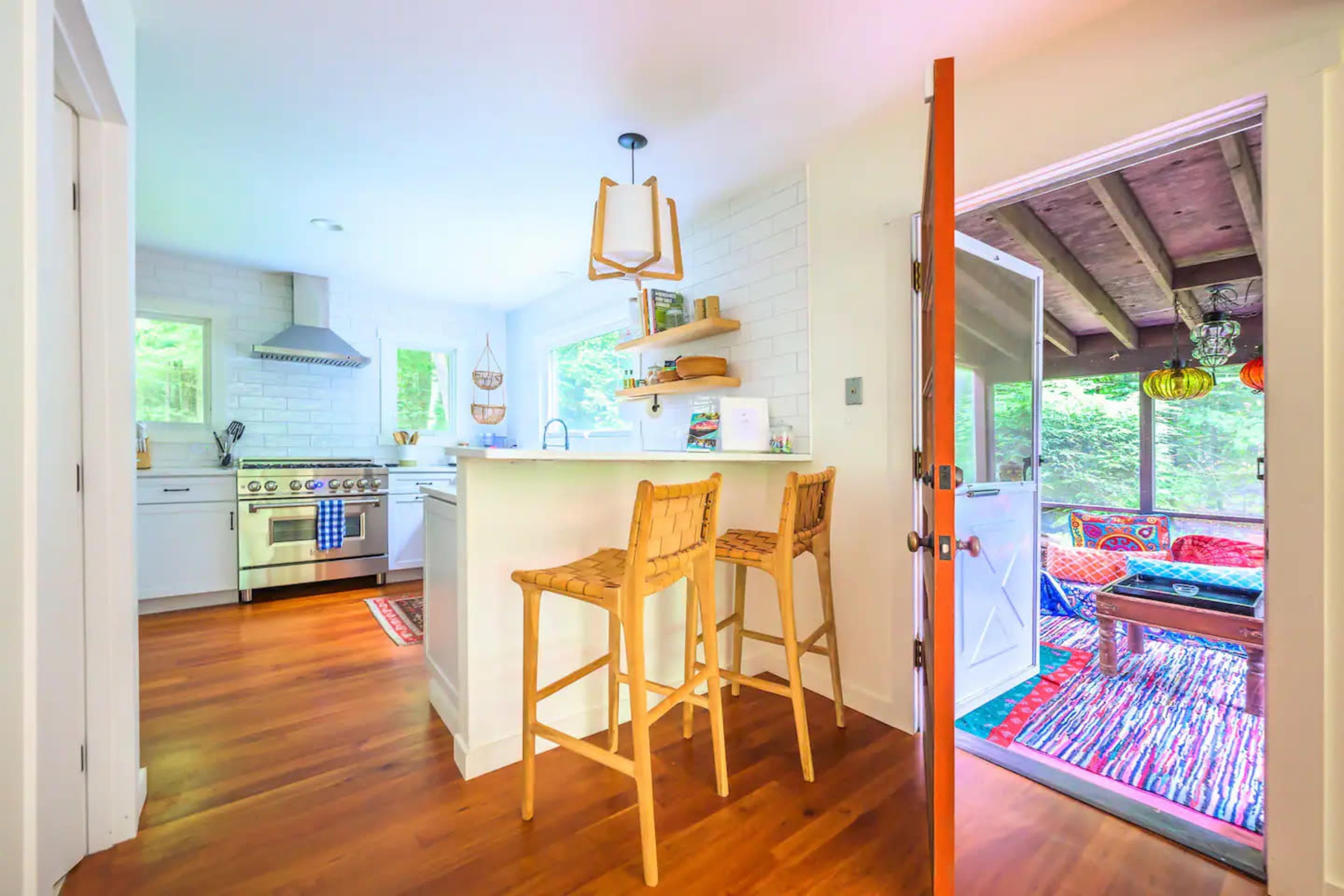A modern kitchen features a white countertop with two tall stools, stainless steel appliances, and an open door leading to a colorful outdoor seating area.