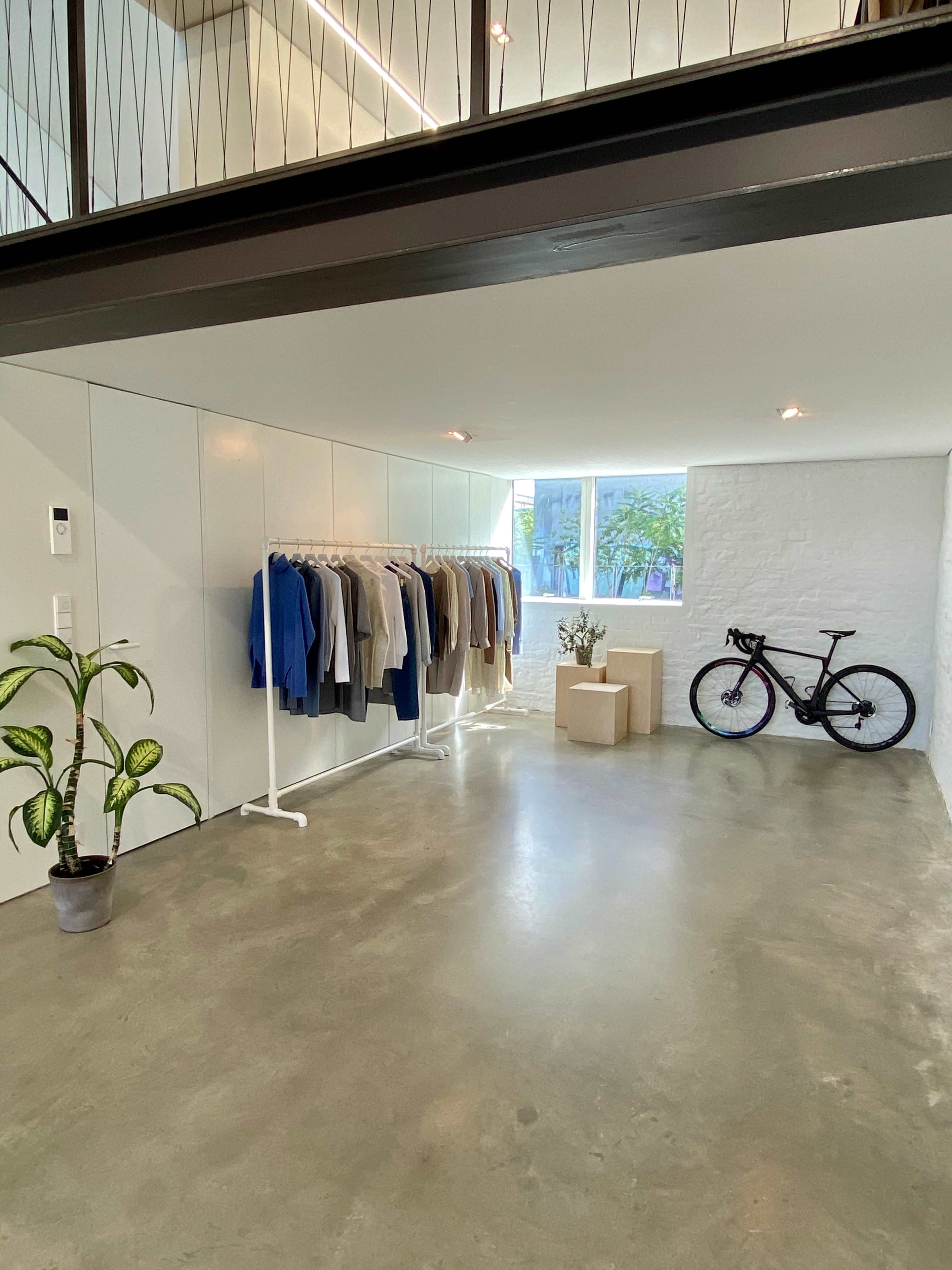 A minimalistic interior space featuring a clothing rack with various garments, a potted plant, a bicycle, and a window allowing natural light.